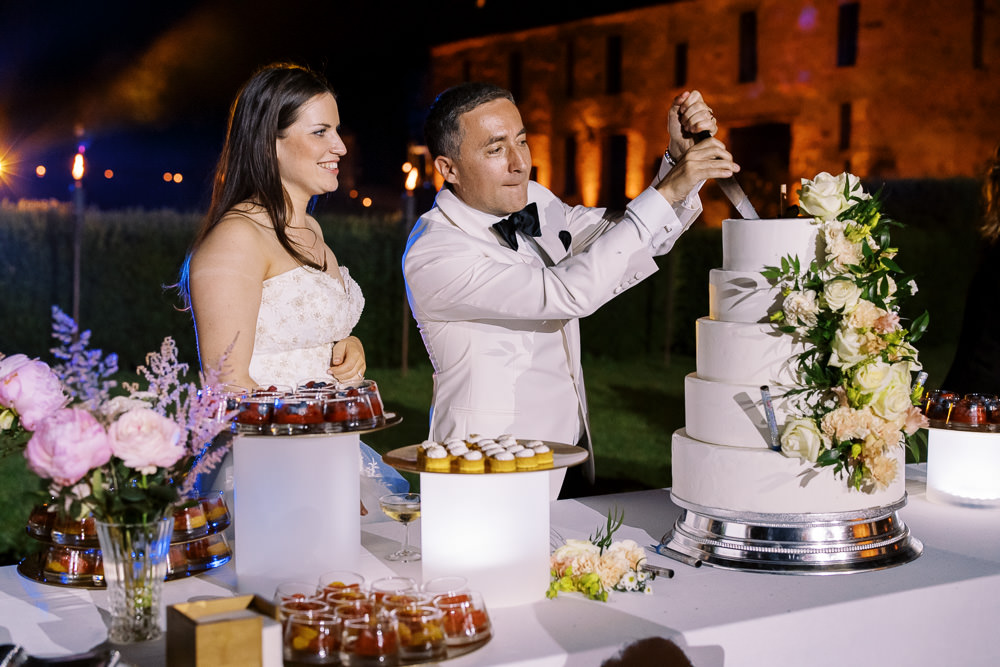 The couple is cutting their wedding cake during an outdoor evening reception, with a warmly lit stone chateau building visible in the background. The bride wears a strapless ivory gown with beaded detailing and stands smiling while the groom, dressed in a white dinner jacket with black bow tie, focuses on pressing the knife into a four-tier smooth white fondant cake decorated with cream and blush roses, garden roses, and greenery on a silver cake stand. The dessert table is covered in a white cloth and displays an extensive spread including individual fruit tart pastries, strawberry verrine cups, small amber-colored desserts in glass jars, and pink peonies with lavender stems in a glass vase as table florals. The shot is a medium portrait-style composition taken at night, with warm uplighting on the venue building and soft glowing lights illuminating the dessert table from below.