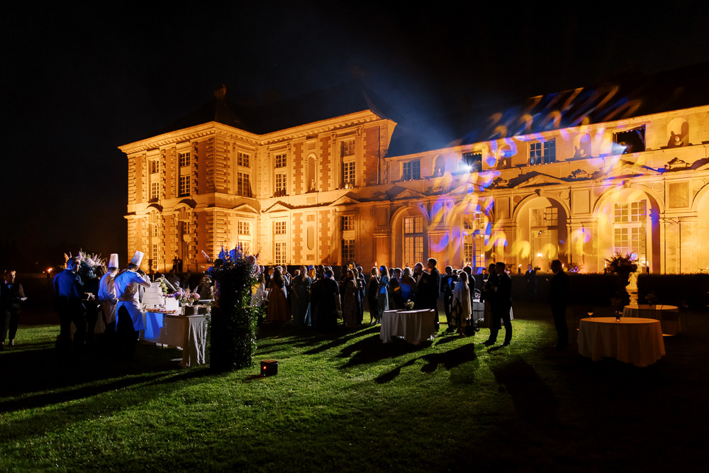 An outdoor evening cocktail hour or reception on the grounds of a large French chateau, captured in a wide shot at night. Approximately 40-50 guests are gathered on the lawn in front of the illuminated chateau facade, which is lit in warm amber and orange tones with blue and purple decorative light projections sweeping across the architecture. A catering station with white linen tablecloths is visible on the left, staffed by a chef in a white toque, and several round cocktail tables with white linens are positioned across the lawn. The chateau is a multi-story classical French building with tall arched windows and formal symmetrical architecture. Potential venue feature image.