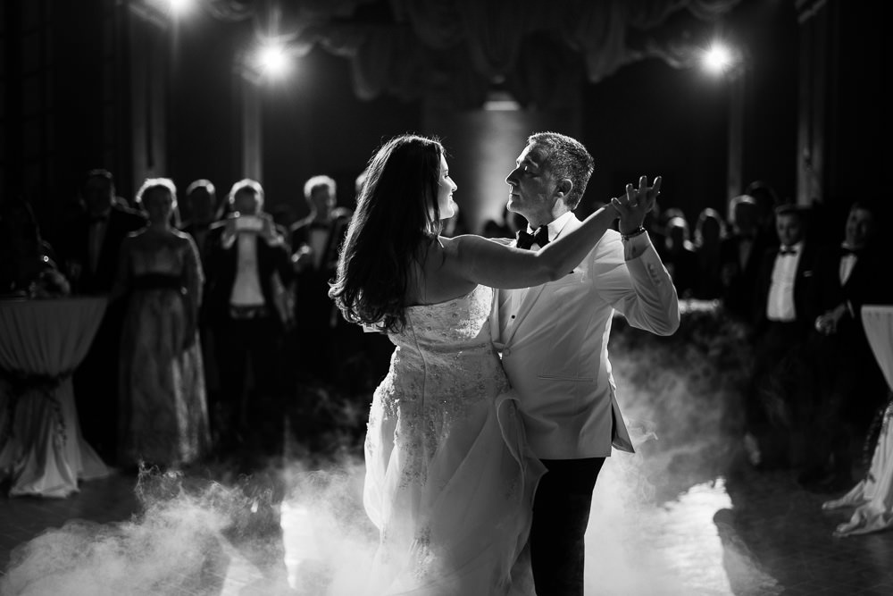 A black-and-white image of a couple sharing their first dance at an indoor reception, captured in a mid-shot from behind and slightly to the side of the bride. The bride wears a strapless lace gown and has long dark hair worn down, while the groom is dressed in a white dinner jacket with a black bow tie and dark trousers. A low-lying fog or dry ice effect rolls across the dance floor around their feet, adding strong contrast against the dark tones of the image. Overhead spotlights illuminate the couple sharply against the darker background, where a crowd of approximately 20–30 formally dressed guests — many in tuxedos and evening gowns — watch from the perimeter of the room. The composition is a portrait-style mid-shot with the couple sharply in focus and the guests rendered in soft blur, emphasizing the formal, classic styling of the event.