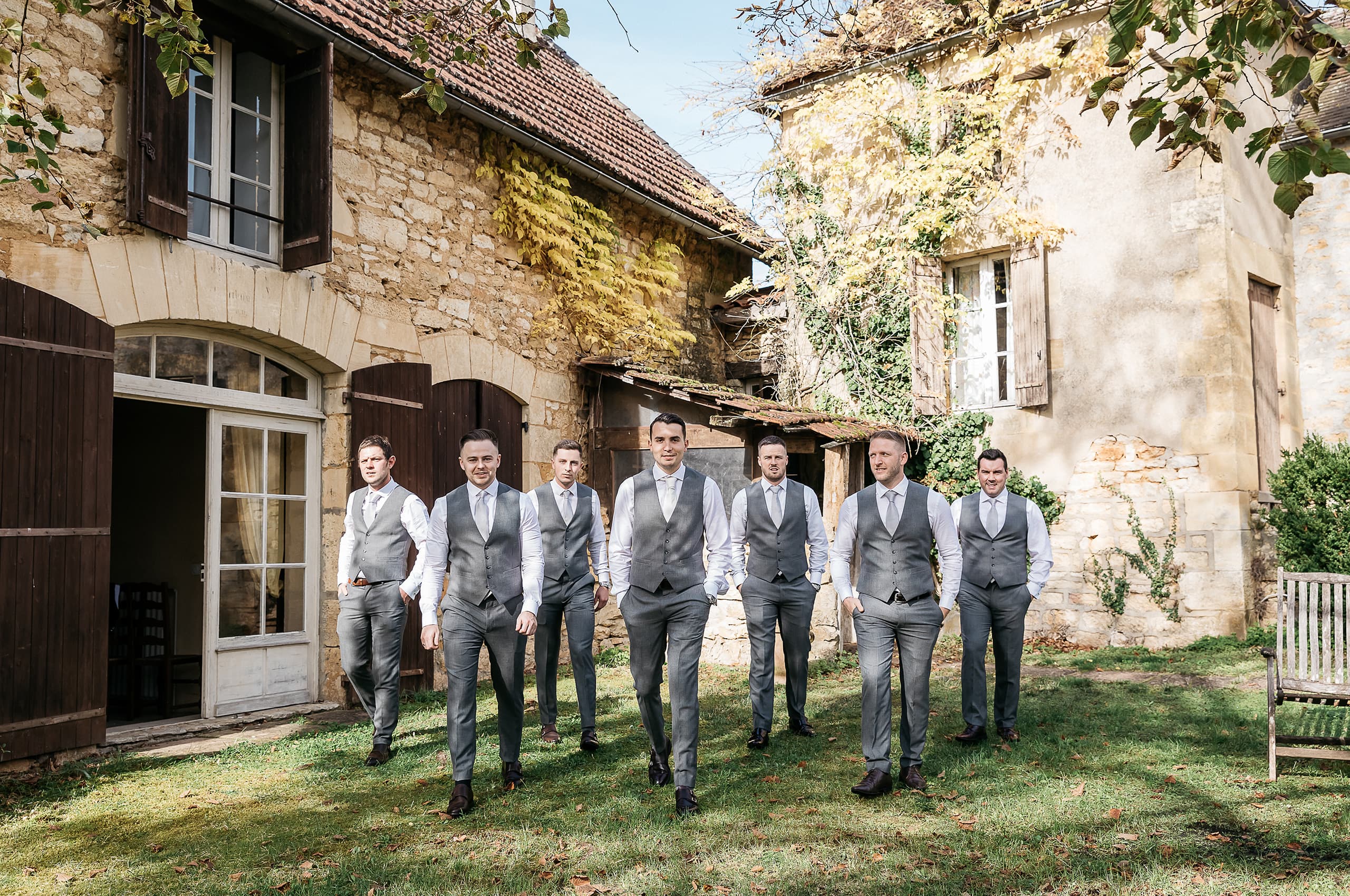 A groomsmen portrait shot outdoors in the courtyard of a rustic French stone-building property, with the group of seven men walking toward the camera in a loose formation. The groom leads at the center, with six groomsmen fanned out behind him. All seven are dressed in matching grey waistcoats, grey trousers, white shirts, and light grey ties, wearing dark leather shoes. The styling is classic and coordinated with a grey palette throughout. The wide-angle shot captures the full-length figures against the backdrop of aged limestone and rendered stone buildings with terracotta tile roofs, wooden shutters, and climbing vines in autumn yellow and green tones.