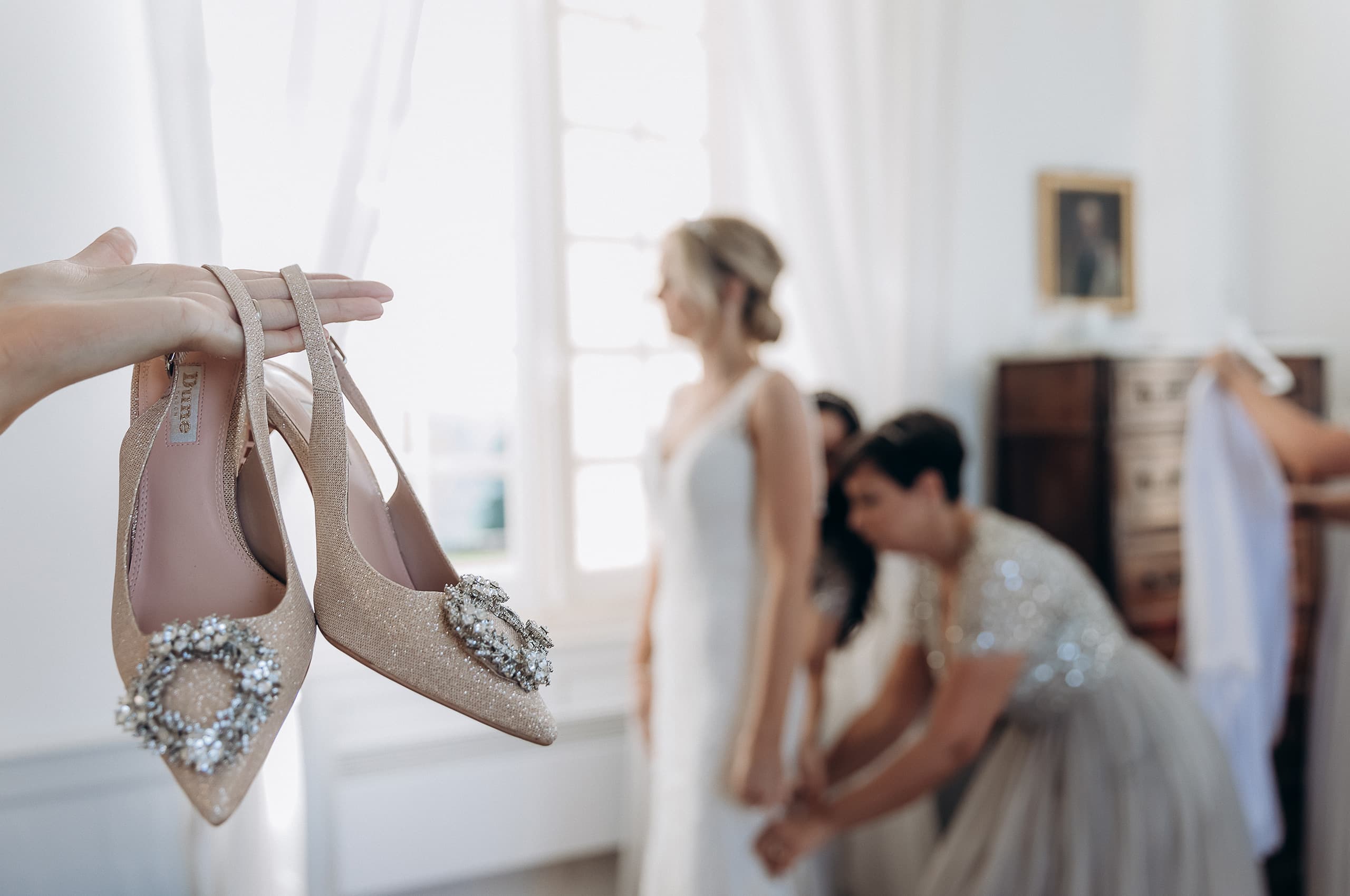 A getting-ready scene shot indoors in a bright, white-walled room that appears to be inside a chateau or manor house, with tall windows, sheer curtains, and a dark wood antique chest of drawers visible in the background. In the foreground, a hand holds up a pair of champagne-gold glitter pointed-toe heels with crystal brooch embellishments and ankle straps, identified by an interior label as Dune. In the soft-focus background, a bride in a fitted ivory sleeveless gown stands while an attendant crouches at her feet, appearing to fasten or adjust something at the hem, and a third person holds what appears to be a white garment near the dresser. The composition uses shallow depth of field to keep the shoes sharp in the foreground while the getting-ready activity behind remains intentionally blurred.