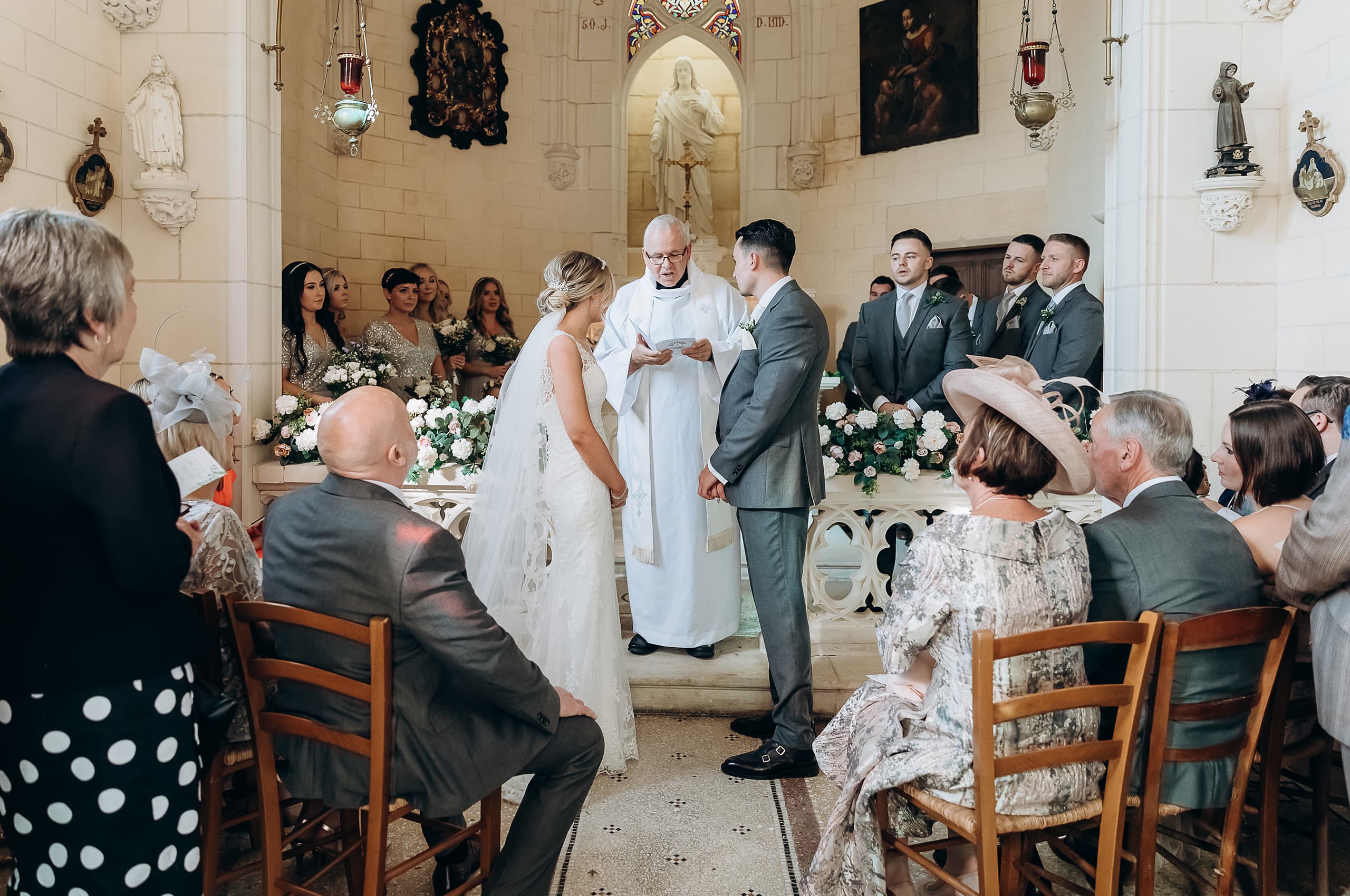 A wedding ceremony taking place inside a small stone chapel with cream-colored limestone walls, religious statues, hanging red lanterns, stained glass, and oil paintings decorating the interior. The bride, in a white lace gown with a veil and hair accessory, stands facing the groom, who wears a light grey suit, while an officiant in white robes reads from a card between them. Three groomsmen in matching grey suits with floral buttonholes stand to the right, and approximately four bridesmaids in silver sequined dresses holding white and blush bouquets are visible to the left. The altar is decorated with arrangements of white hydrangeas, white roses, and green foliage, and seated guests in formal attire fill the wooden chairs on both sides of the aisle. Wide shot taken from the back of the chapel looking toward the altar.