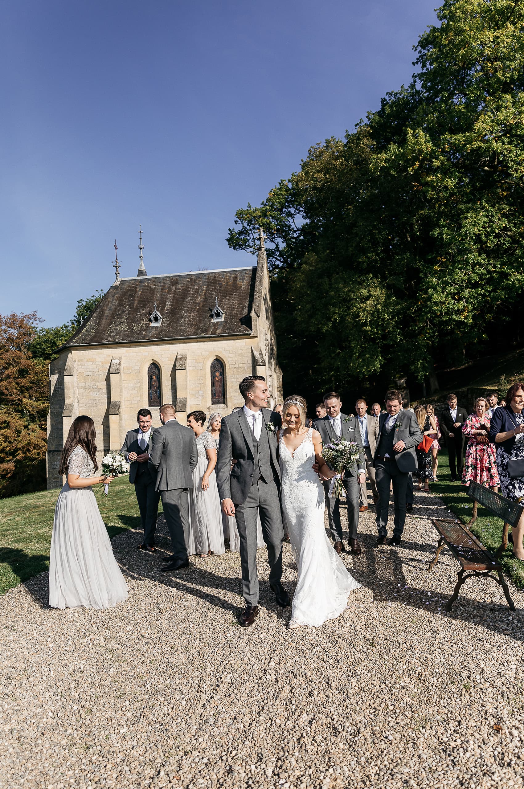 The bride and groom exit a stone chapel following their outdoor ceremony, walking along a gravel path with approximately 20-30 guests, bridal party members, and groomsmen gathered behind and around them. The chapel is a small, historic-looking limestone building with pointed Gothic-style arched windows featuring stained glass, a steep slate roof, and decorative finials. The bride wears a fitted lace wedding dress with a V-neckline and carries a loose bouquet of white and dusty purple blooms, while the groom is dressed in a charcoal grey three-piece suit with a white tie and a white buttonhole flower. Bridesmaids wear floor-length pale silver-grey dresses with embellished tops, and groomsmen are in matching charcoal grey suits. The wide-angle shot captures the full scene in bright natural daylight, with the chapel as a backdrop. Potential venue feature image.