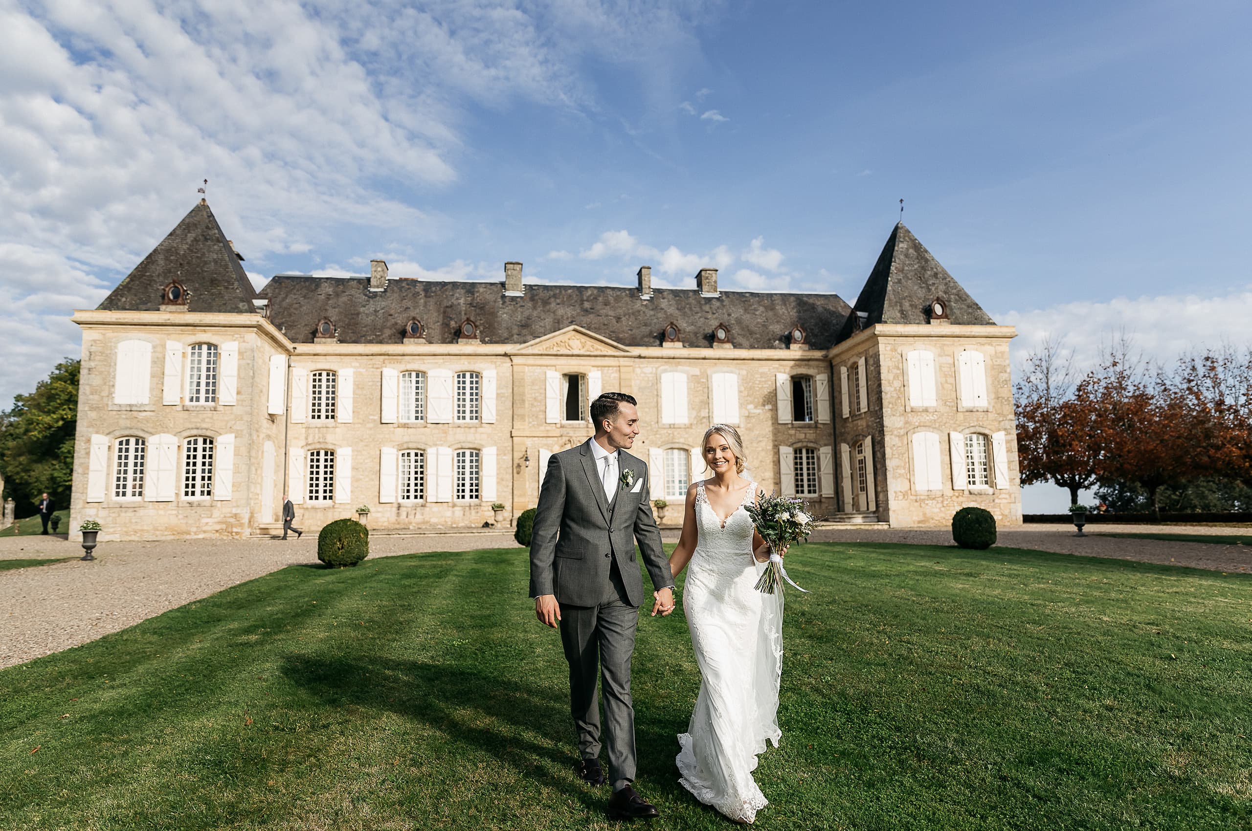 A bride and groom walk hand-in-hand across a manicured lawn in front of a large French chateau with pale stone facades, white shutters, and pointed slate-roofed corner towers. The groom wears a grey three-piece suit with a light grey tie and a white boutonniere, while the bride wears a fitted ivory lace gown with a V-neckline and carries a loose bouquet of white blooms and greenery. The composition is a wide portrait shot that places the couple centrally in the foreground, with the full chateau facade visible behind them and a figure of a guest walking near the entrance in the background. Potential venue feature image.