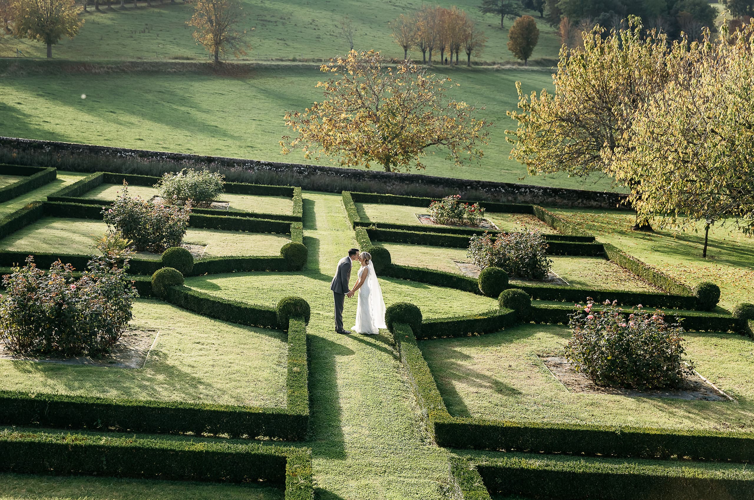 A couple portrait taken from an elevated wide-angle perspective, showing a bride and groom standing at the center of a formal French parterre garden, holding hands and sharing a kiss. The bride wears a white floor-length gown with a veil, and the groom is dressed in a grey suit. The garden features precisely clipped low box hedges arranged in geometric patterns with rounded topiary balls at intervals, and rose bushes with pink blooms planted within the parterre beds. Beyond a low stone boundary wall, open parkland with mature trees showing autumn yellow and orange foliage extends into the distance. The elevated composition emphasizes the scale and symmetry of the formal garden grounds. Potential venue feature image.