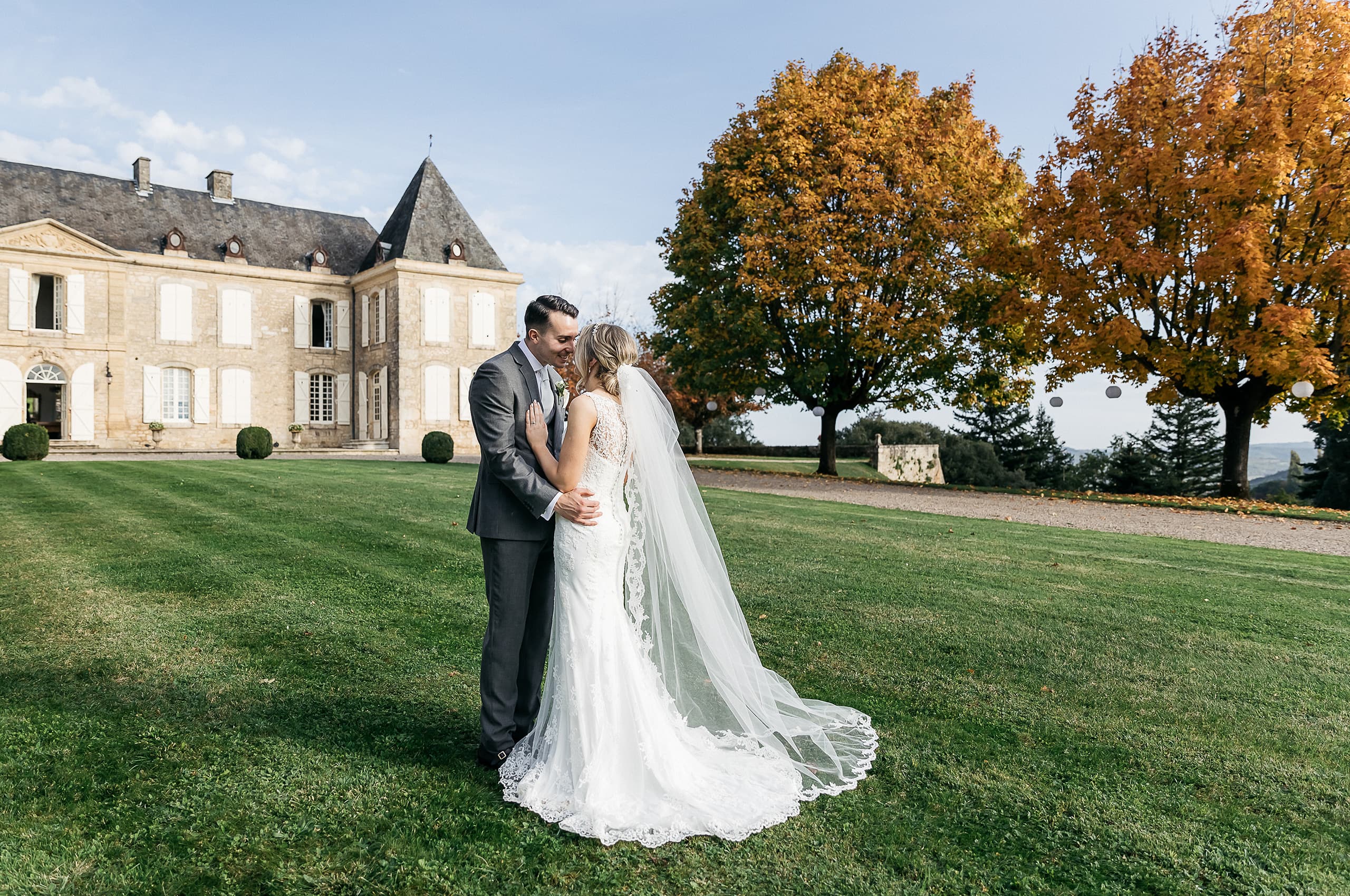 A couple portrait taken outdoors on the grounds of a French chateau during an autumn wedding. The bride wears a fitted ivory lace gown with an open back and a long cathedral-length veil with lace trim that extends across the lawn, while the groom is dressed in a charcoal grey suit with a light grey tie. The two are facing each other and smiling in a relaxed embrace, positioned on a manicured lawn with the multi-story limestone chateau visible to the left and trees with amber and orange autumn foliage to the right. The shot is a wide portrait framing both the couple and the chateau facade in full. Potential venue feature image.
