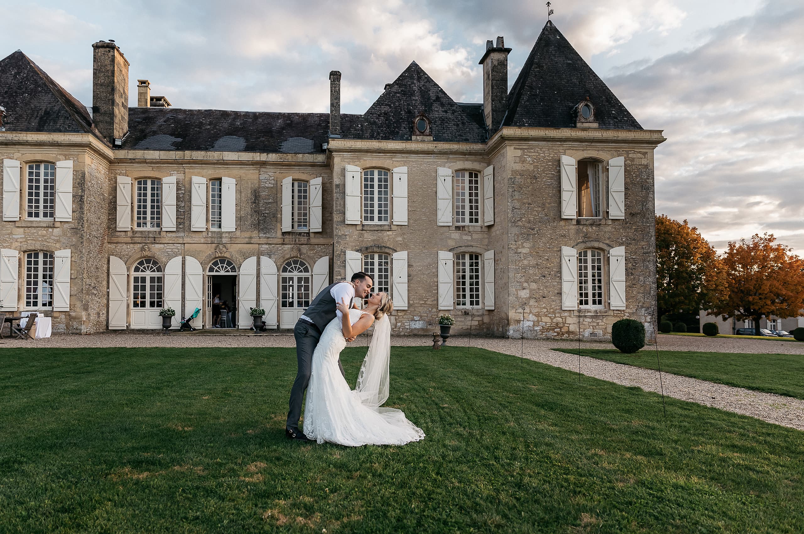 A couple portrait taken outdoors on the lawn of a French chateau, with the groom dipping and kissing the bride in the foreground. The bride wears a fitted lace gown with a long veil, and the groom is dressed in a charcoal grey suit without a jacket, wearing a waistcoat and white shirt. The chateau is a large two-story limestone building with white shutters, arched ground-floor windows, pointed slate turrets, and multiple chimneys, consistent with classic French country architecture. Small potted topiaries and boxwood shrubs are placed along the building's facade, and a gravel pathway runs along the right side of the lawn. The wide-angle shot is taken at dusk with warm ambient light, and a small group of guests can be seen near the open doorway of the chateau in the background. Potential venue feature image.