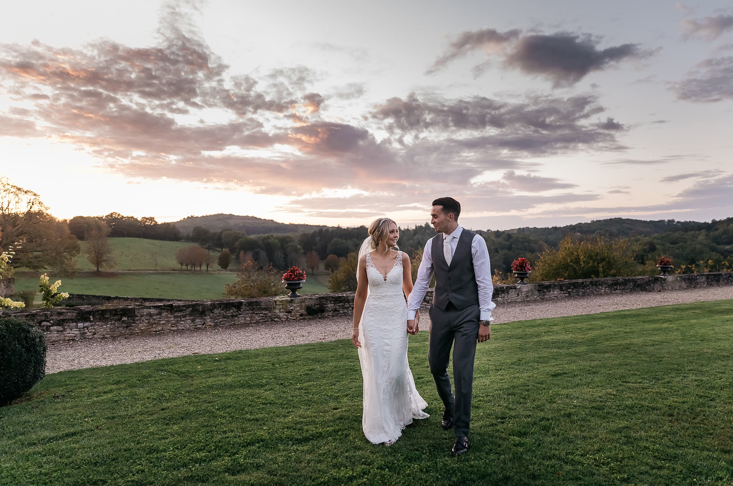 A couple portrait taken outdoors at dusk on the manicured lawn of what appears to be a French chateau or country estate. The bride wears a fitted ivory lace gown with a deep V-neckline and thin straps, while the groom is dressed in a charcoal grey suit with a matching waistcoat, white shirt, and light pink tie; the two walk hand-in-hand looking at each other. A low stone balustrade runs behind them, decorated with dark urn planters holding red floral arrangements, and a gravel pathway is visible beyond it with rolling countryside and wooded hills in the background. The shot is a wide full-length portrait taken at golden hour, with warm ambient light and a wide, dramatic sky providing a natural backdrop.