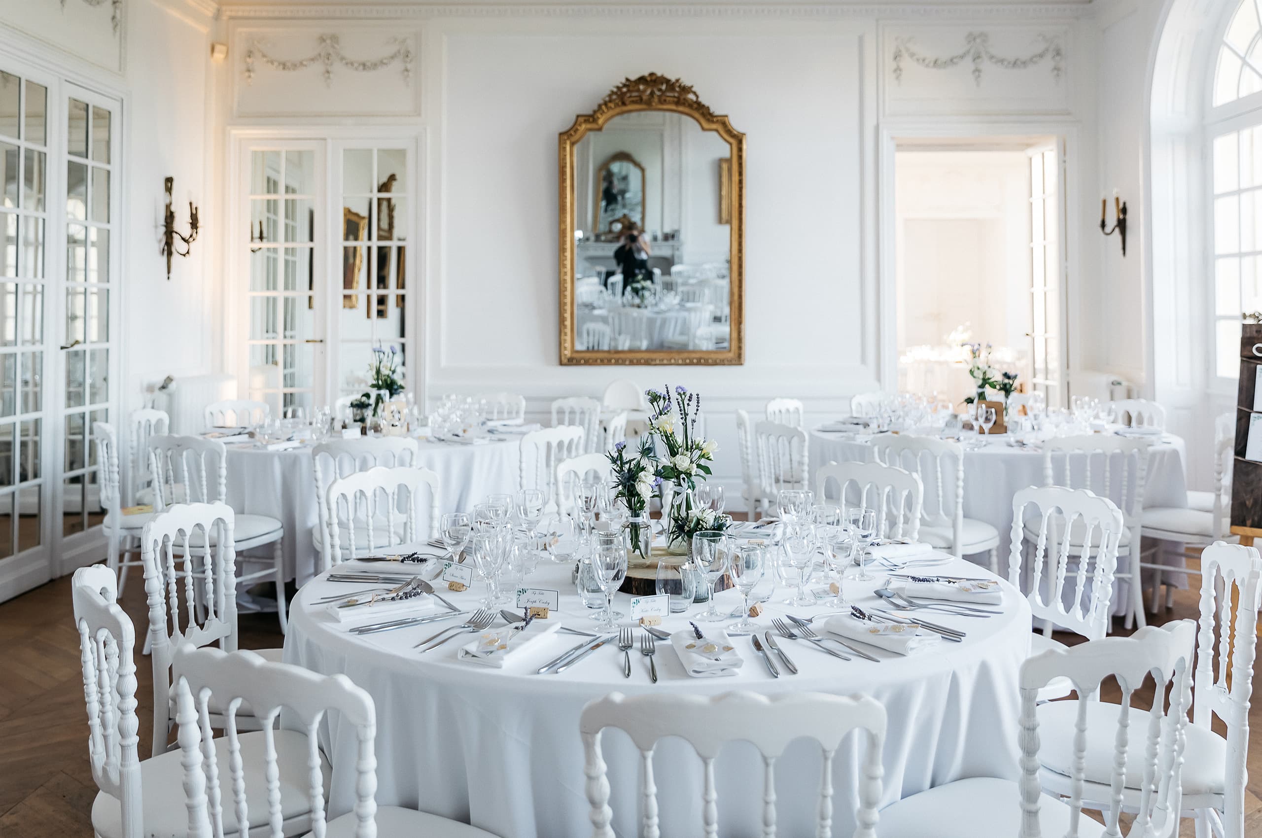 Wide interior shot of a wedding reception room set up for dinner inside what appears to be a French chateau or manor house. The room features white paneled walls with delicate plaster molding friezes, herringbone parquet flooring, ornate gilt-framed mirror above the central wall, bronze wall sconces, and tall French windows flooding the space with natural light. Multiple round tables are dressed in white floor-length linen tablecloths and surrounded by white Napoleon-style spindle chairs, set with silver cutlery, crystal wine glasses, folded white napkins, and small place cards. Each table has a low centerpiece of white roses, lavender, and blue wildflowers in glass vessels, maintaining a white, blue, and soft green palette throughout. The photographer's reflection is visible in the gilt mirror. Potential venue feature image.