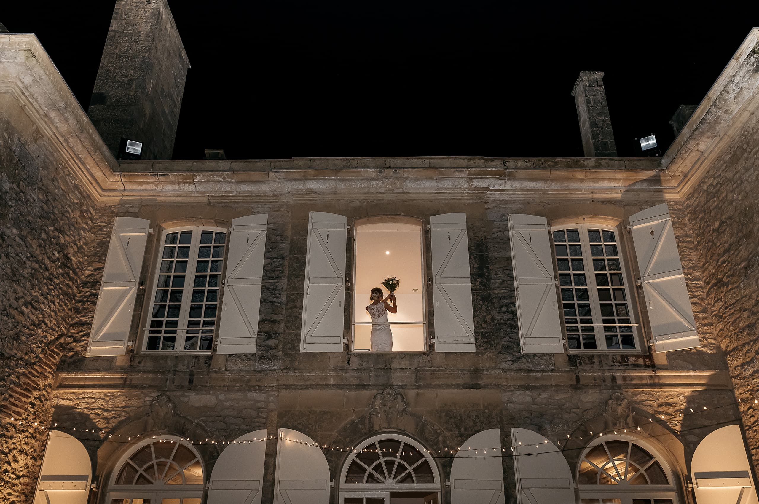 A bride in a fitted white lace-back gown stands in an illuminated upper-floor window of a French chateau at night, holding her bouquet of greenery raised above her head. The wide-angle shot is taken from ground level looking up at the chateau facade, which is warmly lit by exterior uplighting and features white shuttered windows and arched ground-floor openings strung with fairy lights. The contrast between the glowing interior light silhouetting the bride and the deep black night sky creates a dramatic compositional effect. Potential venue feature image.