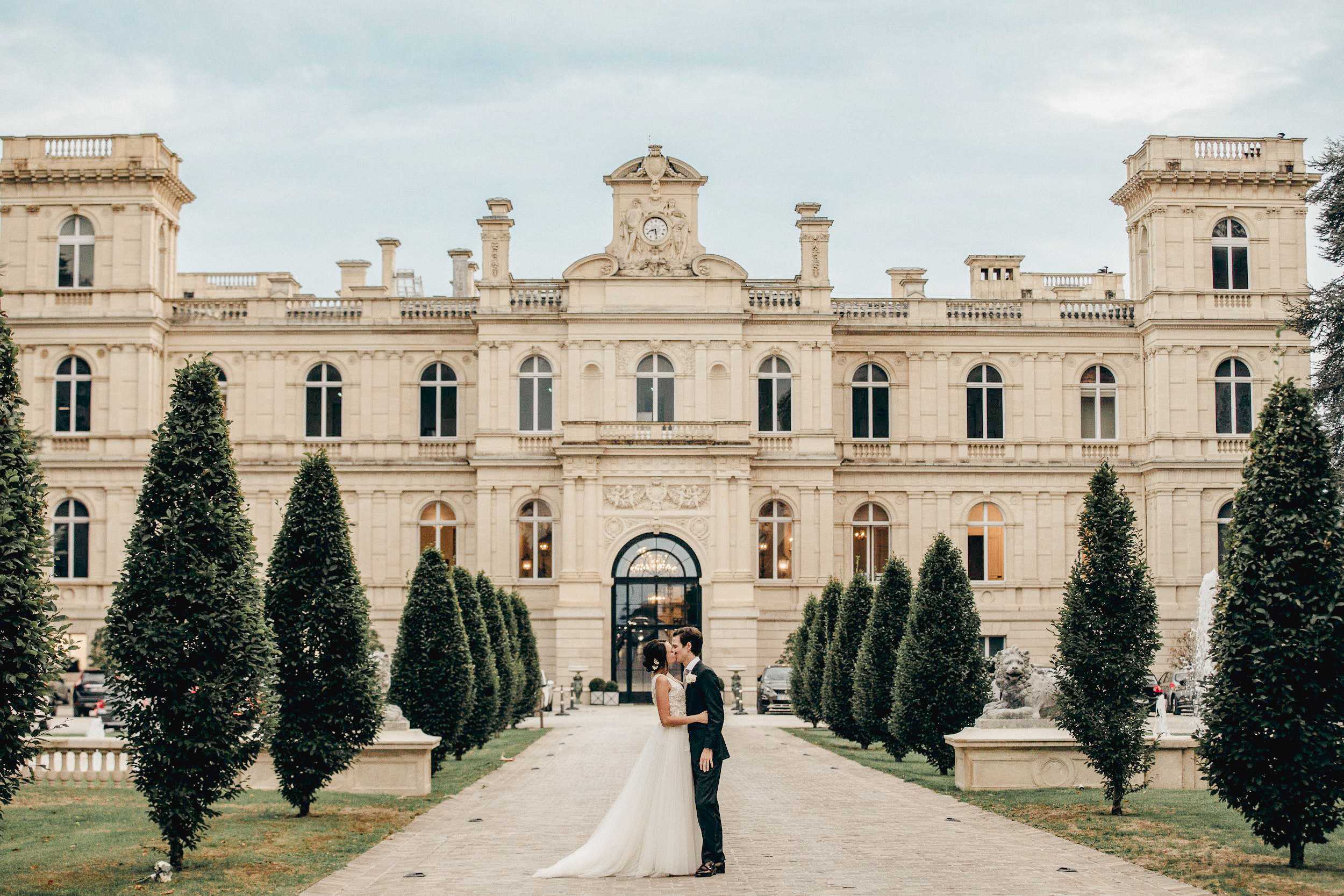 A bride and groom share a kiss on the central paved pathway leading to a grand French chateau, positioned symmetrically in front of the main entrance arch. The bride wears a sleeveless ballgown with an embellished lace bodice and a full ivory tulle skirt with a train, while the groom is dressed in a dark navy suit. The chateau facade is a multi-story classical French architecture building in cream stone, featuring arched windows, ornate pediment with a clock, sculpted friezes, and stone lion statues flanking the entrance; the interior glows warmly through the ground-floor windows. The pathway is lined on both sides with tall, narrow columnar topiary trees planted in formal rows, and a fountain is partially visible to the right. This is a wide-shot couple portrait taken at ground level with the couple centered against the full facade of the building. Potential venue feature image.