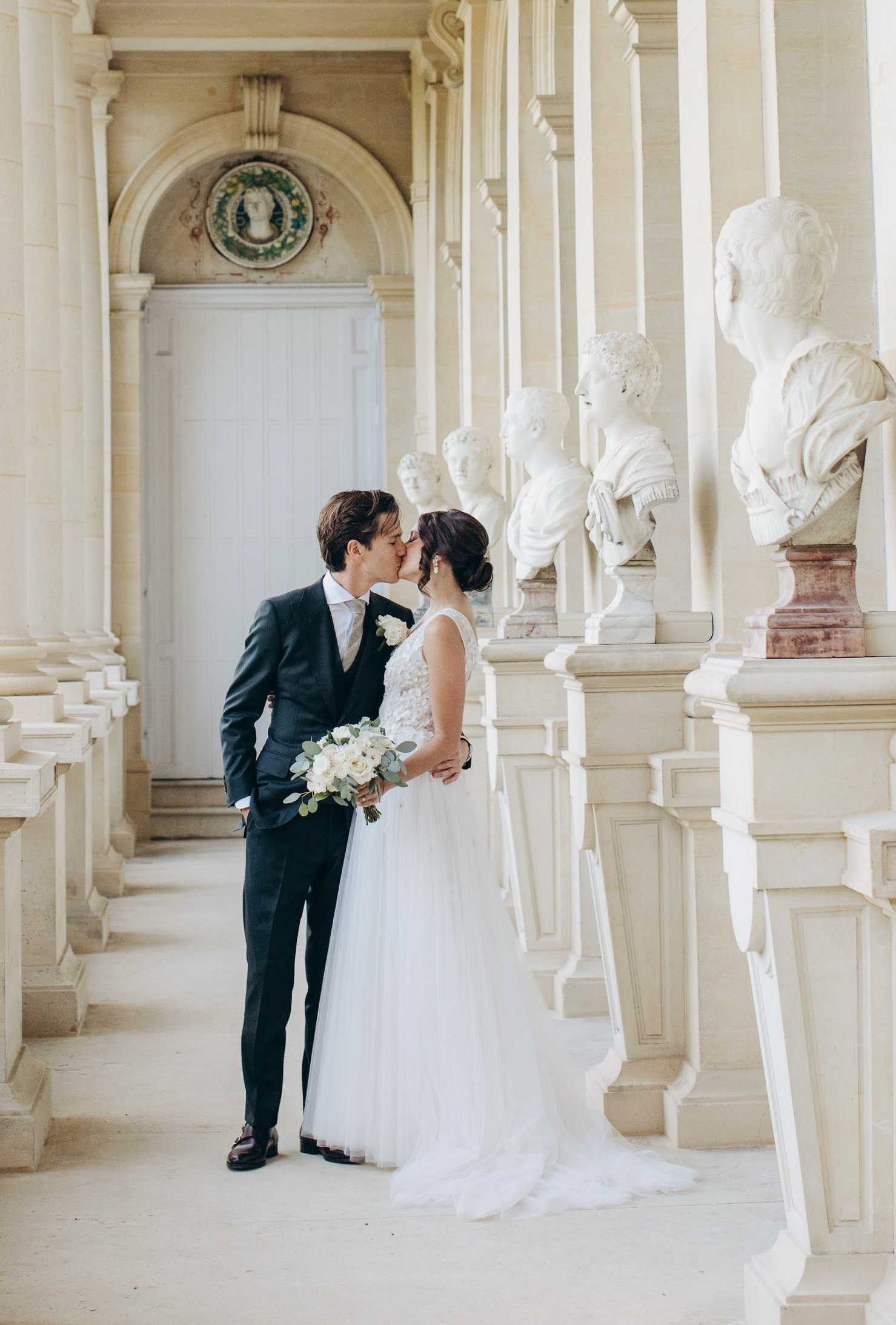 A couple portrait taken in a grand covered colonnade lined with white marble portrait busts on pedestals, consistent with a classical French château or palace gallery. The bride wears a sleeveless ivory tulle ball gown with a lace-embroidered bodice and a trailing skirt, her dark hair styled in an updo; she holds a bouquet of white garden roses and eucalyptus. The groom wears a charcoal three-piece suit with a light taupe tie and a white rose boutonnière. The two are kissing in the center of the frame, with the colonnade receding symmetrically behind them toward an arched doorway decorated with a painted medallion. The shot is a full-length portrait with a classic, symmetrical composition.