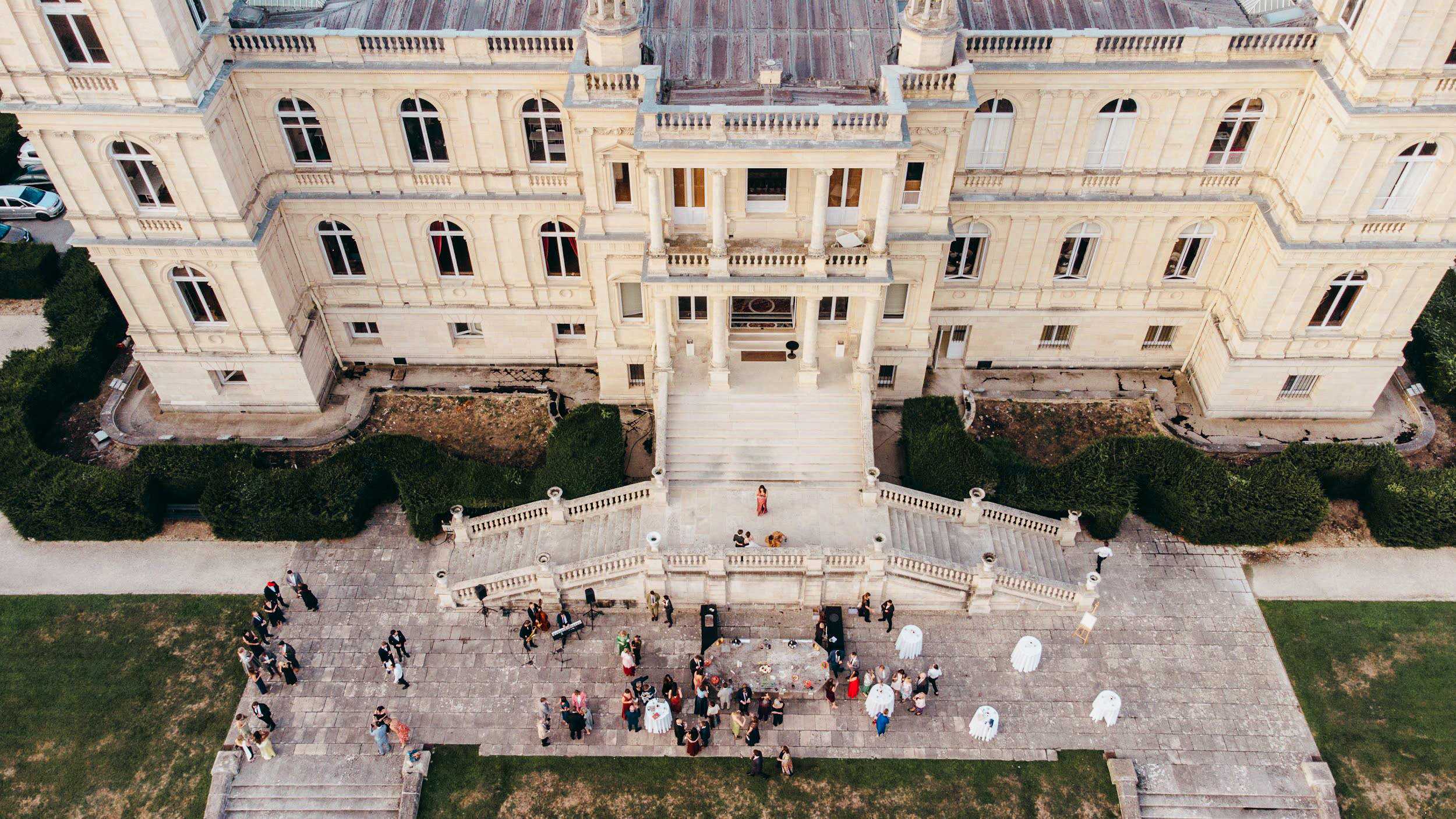 An aerial drone shot of a cocktail hour taking place on the stone terrace and forecourt of a large French chateau with cream-colored neoclassical architecture, featuring arched windows, balustrades, and a grand central staircase with double flights. Approximately 50 to 60 guests are mingling across the paved terrace in front of the building, dressed in formal attire in a variety of colors. A live musician with a keyboard setup is visible on the left side of the terrace, and several round high-top cocktail tables dressed in white linen are scattered across the space, along with what appear to be tall white floral or balloon installations on the right side. One figure in a red or colorful dress stands prominently on the upper landing at the base of the main entrance stairs, possibly a performer or the bride. The shot is taken at or near golden hour, casting warm light across the pale stone facade. Potential venue feature image.