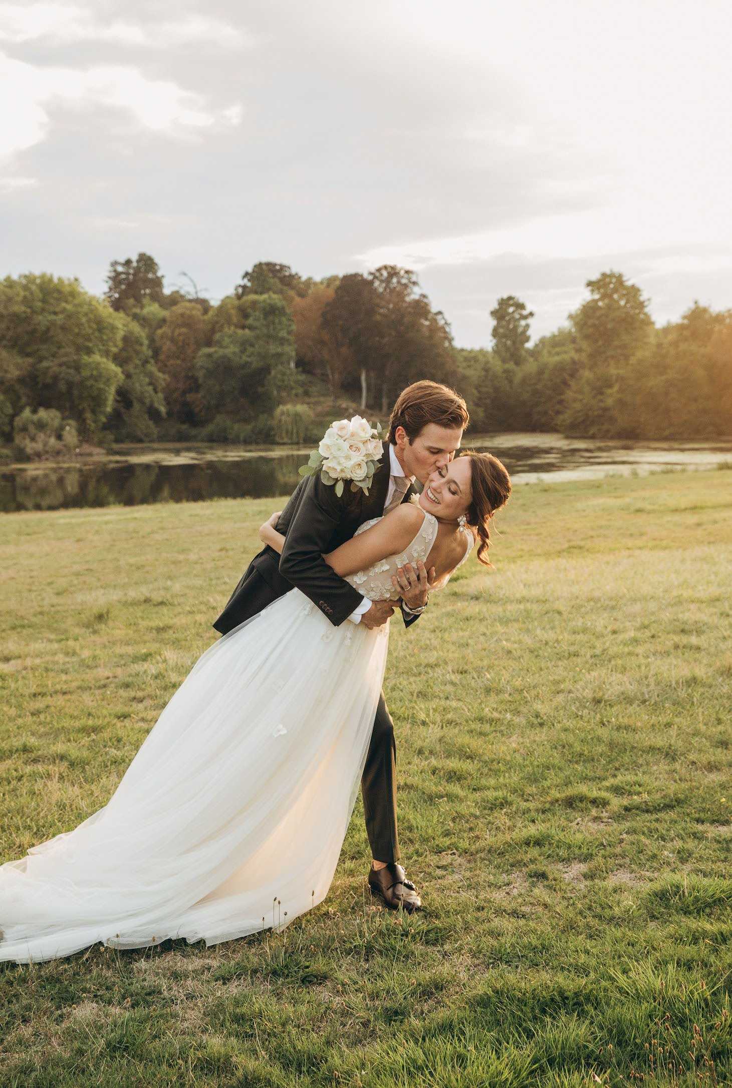 Outdoor couple portrait taken in a wide open meadow with a calm pond and tree line in the background, shot during golden hour with warm directional light. The groom, dressed in a dark navy suit with a light tan tie, is dipping and kissing the bride on the cheek while she laughs, holding her bouquet of ivory roses and soft greenery behind his back. The bride wears a fitted floral-appliqué bodice wedding gown with a full ivory tulle skirt and a long train that fans across the grass. The composition is a full-length portrait with a slightly wide angle, capturing both figures against the open landscape.