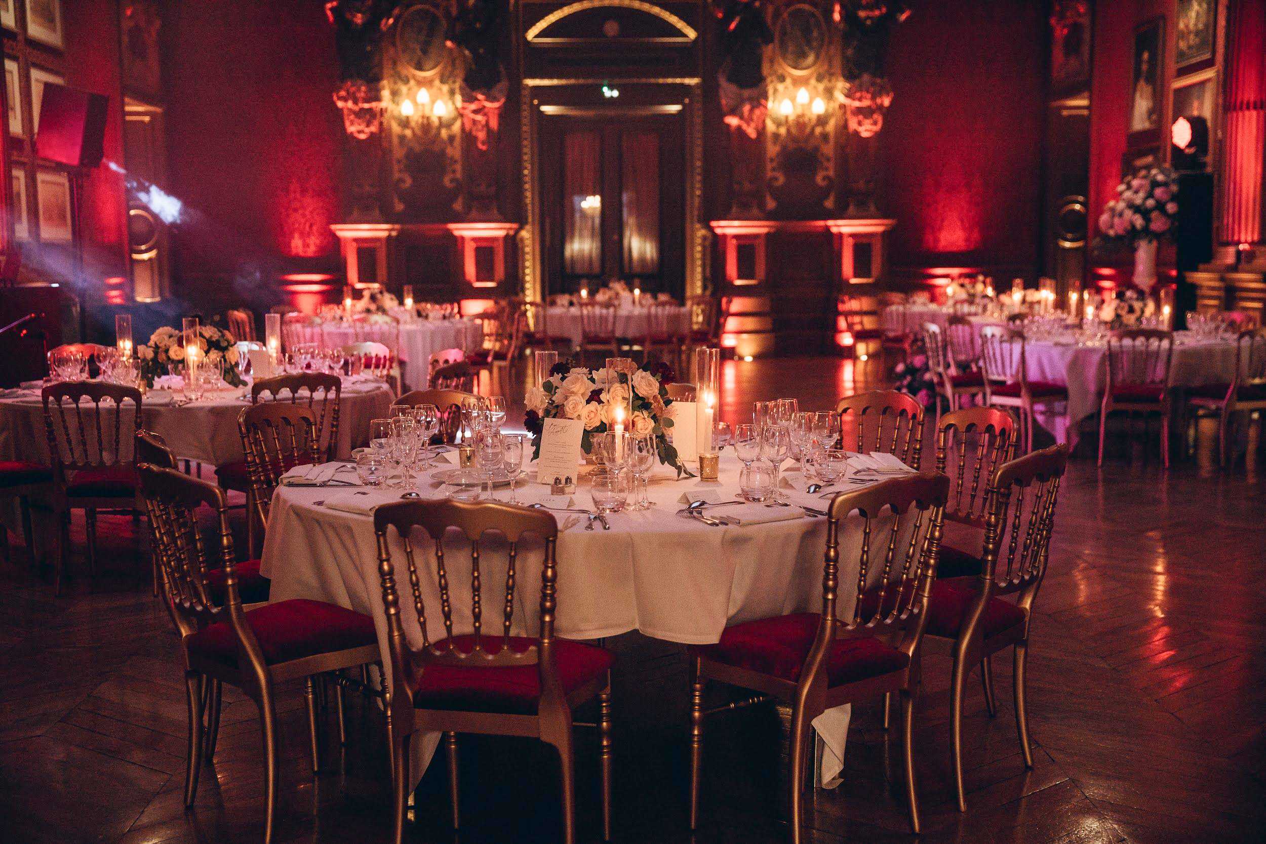 Wedding reception setup inside a grand ballroom with deep red walls, ornate gold moldings, gilt-framed paintings, and chandelier wall sconces casting warm amber light. The room is filled with multiple round tables dressed in white linen, surrounded by gold Napoleon-style chairs with red upholstered seat cushions. The foreground table centerpiece features white roses with dark burgundy accents and greenery, flanked by tall glass cylinder candles and small votive candles, with full crystal glassware and silver cutlery place settings visible. Red uplighting washes the walls throughout the room, reinforcing a classic, formal color palette of red, gold, and white. Wide-angle interior shot taken before guests are seated. Potential venue feature image.