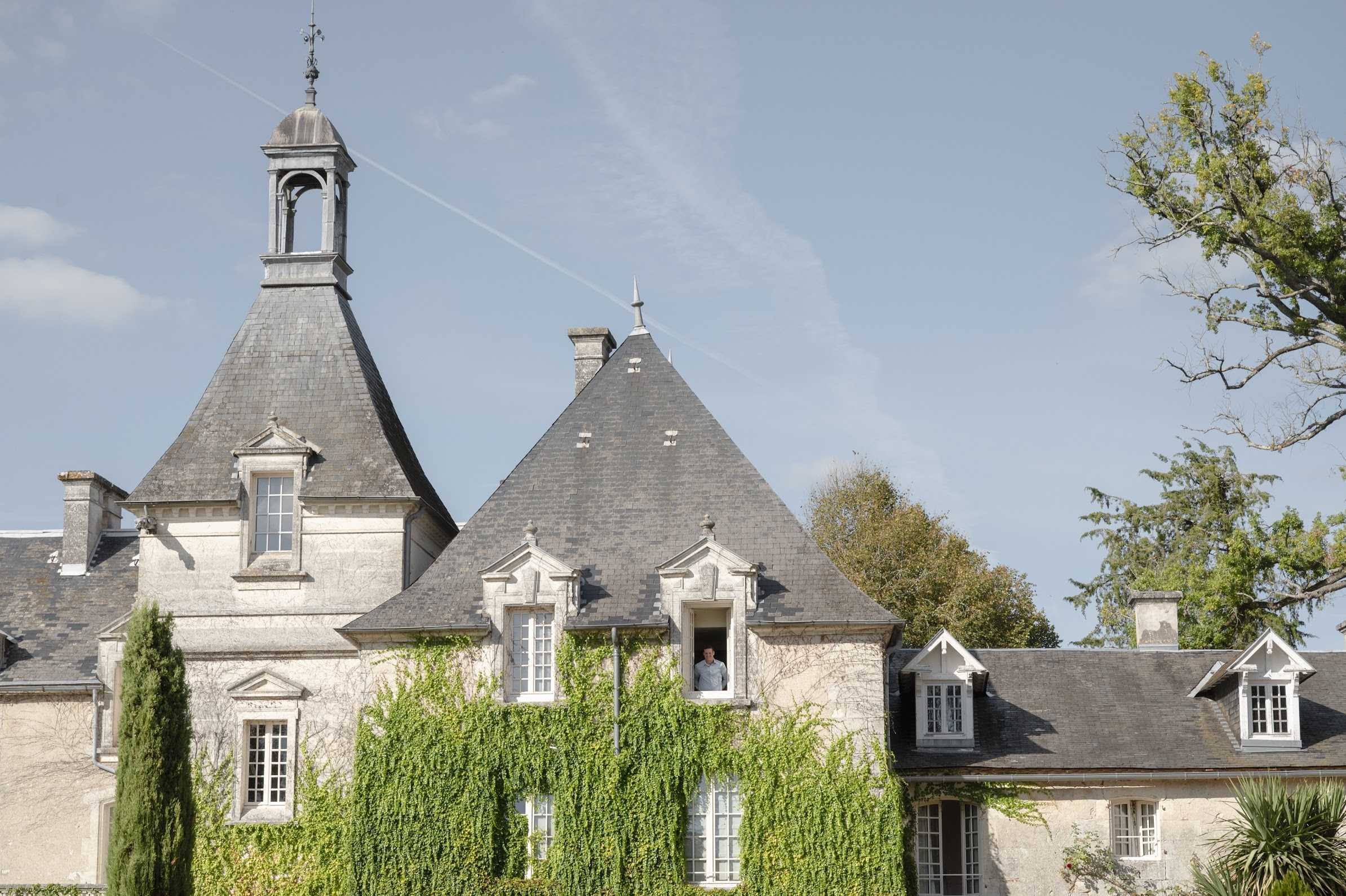 A wide exterior shot of a classic French chateau with pale limestone facades, slate grey conical turret roofs, and ornate dormer windows. The building is partially covered in green ivy climbing the upper stories, and a single person — likely the groom or a guest — leans out of an open upper-floor window. The architecture features a prominent bell-topped tower, pointed spires, and traditional French Renaissance-style detailing. Potential venue feature image.