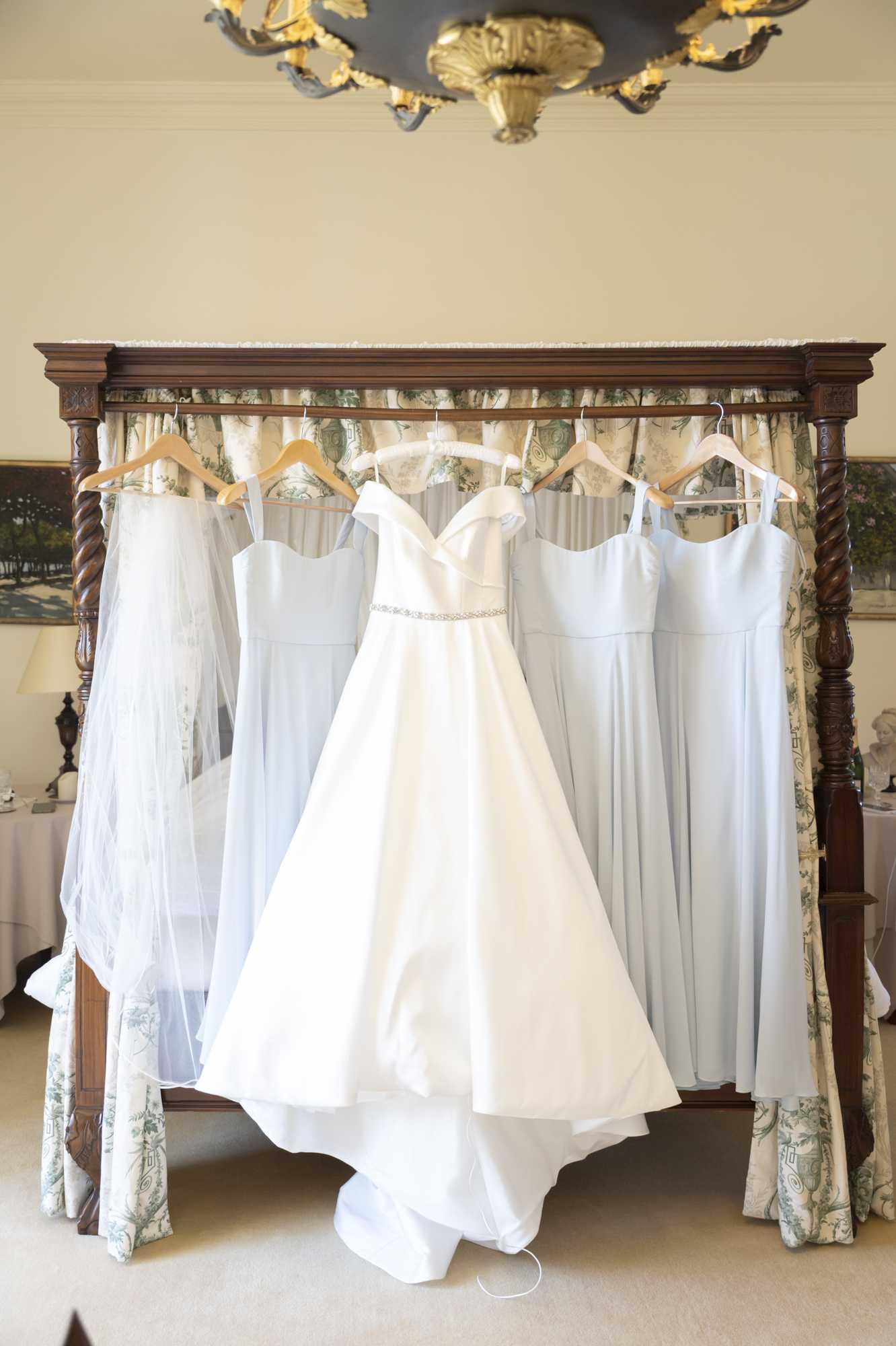 A getting-ready detail shot showing a white off-the-shoulder A-line bridal gown with a slim crystal-embellished waistband hanging from the headboard of an ornate dark wood four-poster bed, flanked by four pale ice-blue spaghetti-strap bridesmaid dresses and a sheer cathedral-length veil, all on wooden hangers. The bed features floral toile canopy curtains in green and cream, and a gilded ornamental chandelier is partially visible at the top of the frame. The setting is a classic, well-appointed interior room with cream walls, suggesting a chateau or manor house bridal suite. The composition is a wide, centered shot designed to display all the dresses together.