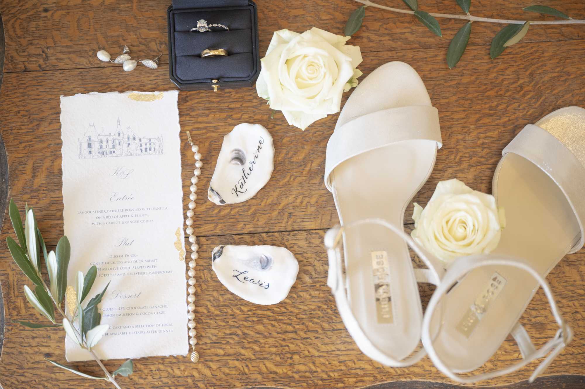 A flat lay of wedding day details arranged on a warm wooden surface, featuring a deckle-edged menu card printed with a chateau illustration and the initials 'K&J' alongside a three-course French menu with gold accent details. Two oyster shells are used as place cards with the names 'Katherine' and 'Lewis' written in calligraphy, and a pair of white strappy block-heel sandals sits alongside two cream roses. A black velvet ring box holds a diamond engagement ring and a plain gold band, accompanied by small pearl drop earrings and a pearl bracelet or necklace. Olive branch sprigs with small white flowers are placed at the lower left corner, completing the classic, French-countryside-inspired styling.