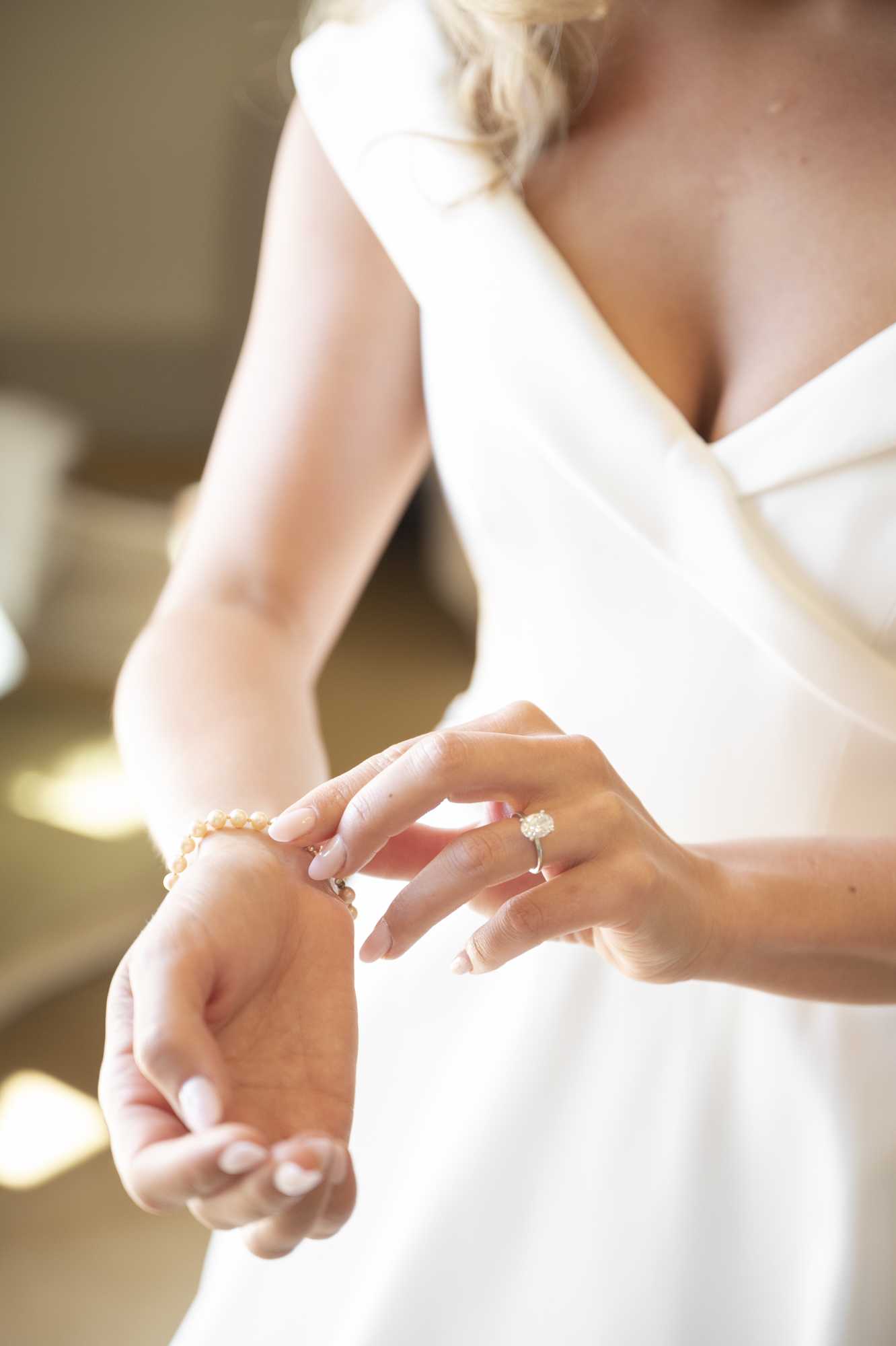 A close-up detail shot capturing a bride fastening a delicate pearl bracelet around her wrist during the getting-ready portion of the day. She is wearing a white dress with a V-neckline and structured bodice, and her engagement ring — a solitaire oval diamond on a silver band — is clearly visible on her left hand. Her nails are painted a soft nude-pink, and her blonde hair is loosely pinned up. The background is softly blurred with warm, diffused indoor light.