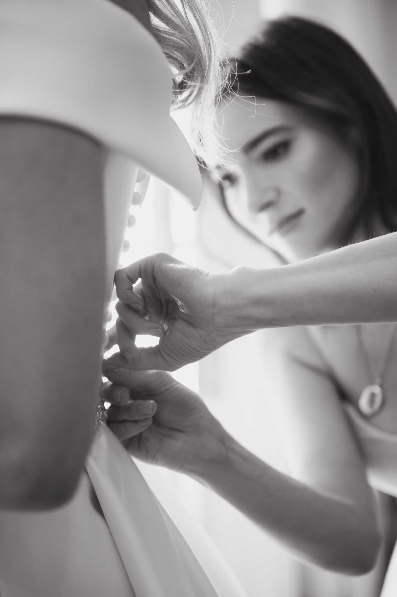 Black-and-white close-up shot taken during the getting-ready portion of a wedding, showing a bride having the buttons or fastening on the back of her wedding dress secured by a second person whose hands are visible in the foreground. The bride, a dark-haired woman with an updo with loose strands, looks downward with a focused expression, wearing a strapless or off-shoulder gown; a small circular pendant necklace is visible at her collarbone. The composition uses a shallow depth of field, with the hands and dress fastening sharp in the foreground and the bride's face softly blurred in the background, creating a layered, intimate perspective. Bright, soft window light illuminates the scene, producing gentle highlights and mid-range contrast typical of black-and-white photography.