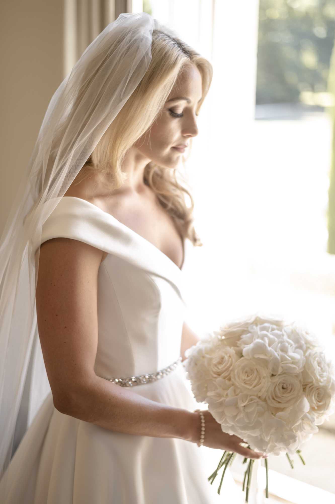 A bridal portrait taken indoors, with the bride standing in profile near a large window with strong natural backlight. She wears an ivory off-the-shoulder satin gown with a crystal and pearl embellished waistband, paired with a full-length plain-edged veil worn over loose blonde hair. She holds a tightly packed round bouquet composed entirely of ivory and white roses and white hydrangeas, and wears a pearl bracelet. She is looking downward with eyes closed. The composition is a medium close-up portrait shot from the side, with the bright window creating a high-key, softly overexposed background.