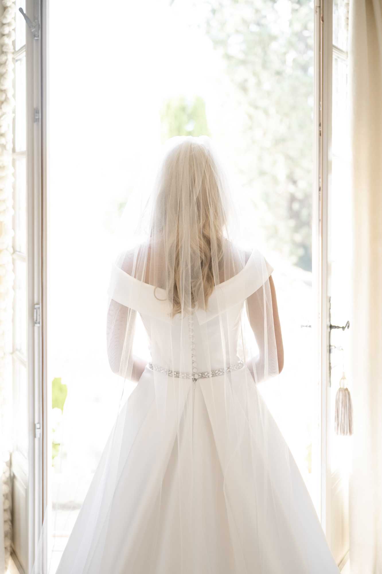 A portrait shot of a bride photographed from behind, standing in an open set of tall French doors that flood the frame with bright natural backlight. She is wearing an ivory off-the-shoulder ballgown with a corseted back closure and a thin crystal or beaded belt at the waist, paired with an elbow-length tulle veil over loosely curled blonde hair. The interior setting appears to be a chateau or manor house room, with cream-painted woodwork, draped curtains, and a decorative tassel key visible on the door. The bright overexposed exterior suggests a garden or grounds beyond the doorway.