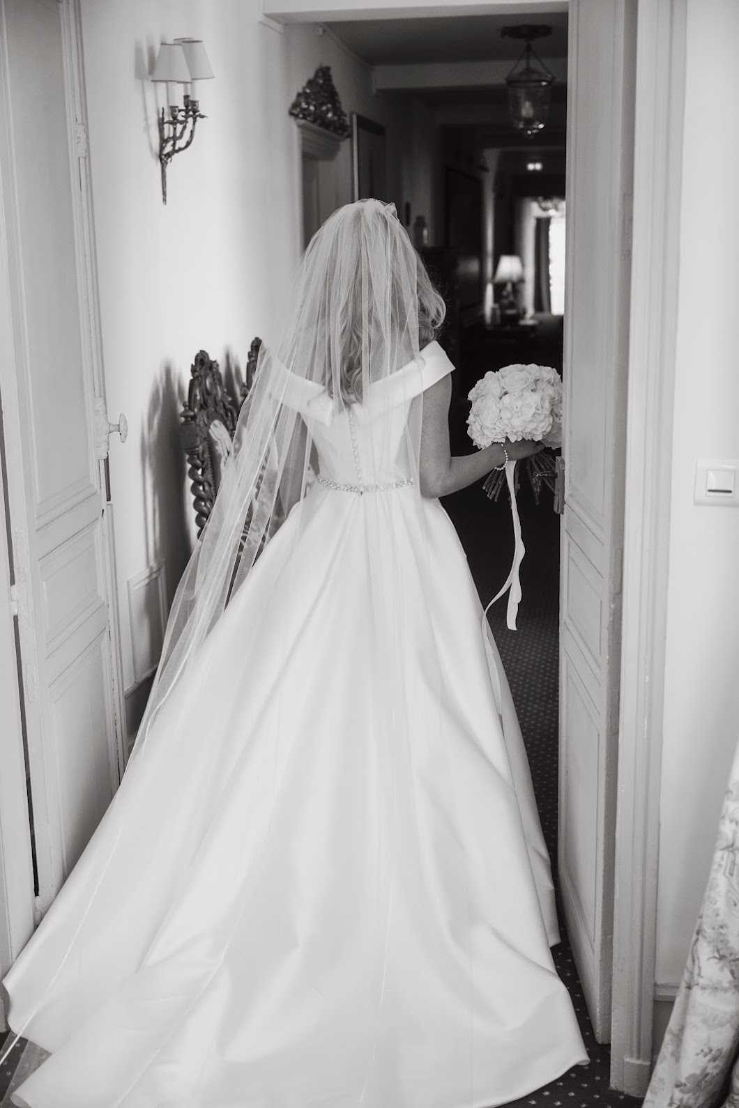 This black-and-white getting-ready or departure shot captures a bride photographed from behind as she stands in a doorway, transitioning between two interior rooms. She is wearing an off-the-shoulder ball gown with a structured bodice, a delicate embellished waist belt, and a full skirt with a substantial train spread across the floor. A mid-length tulle veil falls from her upswept blonde hair. In her right hand she holds a round bouquet of what appear to be white peonies or roses with ribbon streamers. The interior setting features classic French architectural details including paneled white doors, an ornate gilded wall sconce with lamp shades, and a carved dark wood headboard visible to the left, suggesting a chateau or upscale hotel bridal suite. The image is a full-length portrait shot with soft, even tones and moderate contrast typical of B&W film-style processing.