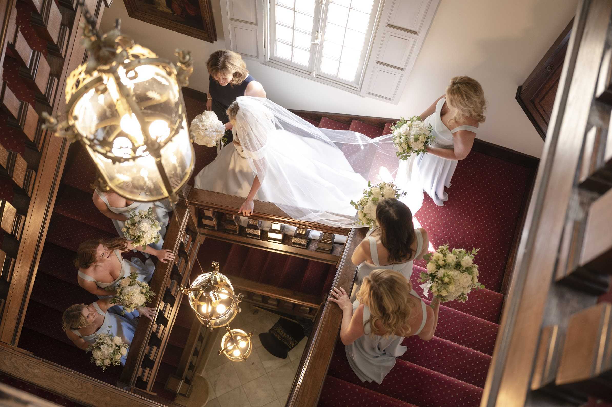 An overhead wide shot captures a bride descending a grand interior staircase with a long cathedral-length veil and a full white ballgown, accompanied by approximately five bridesmaids in pale powder blue dresses and one woman in a navy dress who appears to be assisting with the veil and train. The staircase features dark wood banisters, deep red carpet with a polka-dot pattern, and is lit by two ornate brass and glass lantern-style pendant lights hanging in the stairwell. Each bridesmaid carries a bouquet of ivory roses and white blooms with eucalyptus greenery, while the bride holds a compact all-white bouquet. A large framed oil painting is partially visible on the upper landing wall, reinforcing the classic, formal interior setting of what appears to be a chateau or manor house. The composition is shot from directly above, giving a dramatic bird's-eye view of the processional descent.
