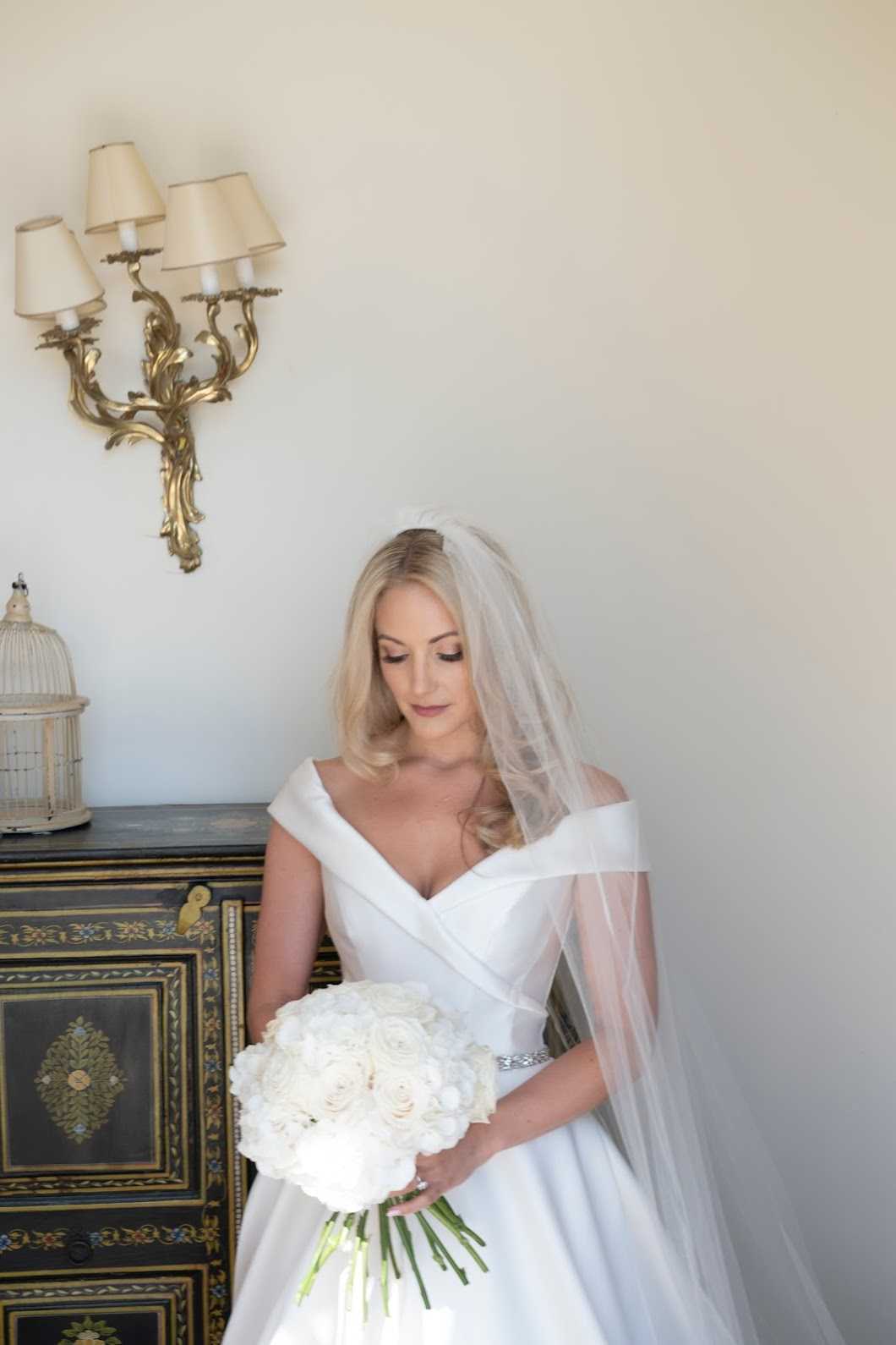 A bridal portrait taken indoors, showing the bride standing and looking downward at her bouquet of tightly packed ivory roses with exposed green stems. She wears an off-the-shoulder white satin ball gown with a crystal-embellished waist belt and a plain-edge tulle veil worn at mid-length. Her blonde hair is worn down in soft waves. The setting features a cream wall with an ornate gold candelabra-style wall sconce, and a dark painted wooden chest of drawers with floral inlay decoration and a small decorative birdcage on top. The overall styling is classic and formal. This is a medium portrait shot with the decorative furniture providing context on the left side of the frame.