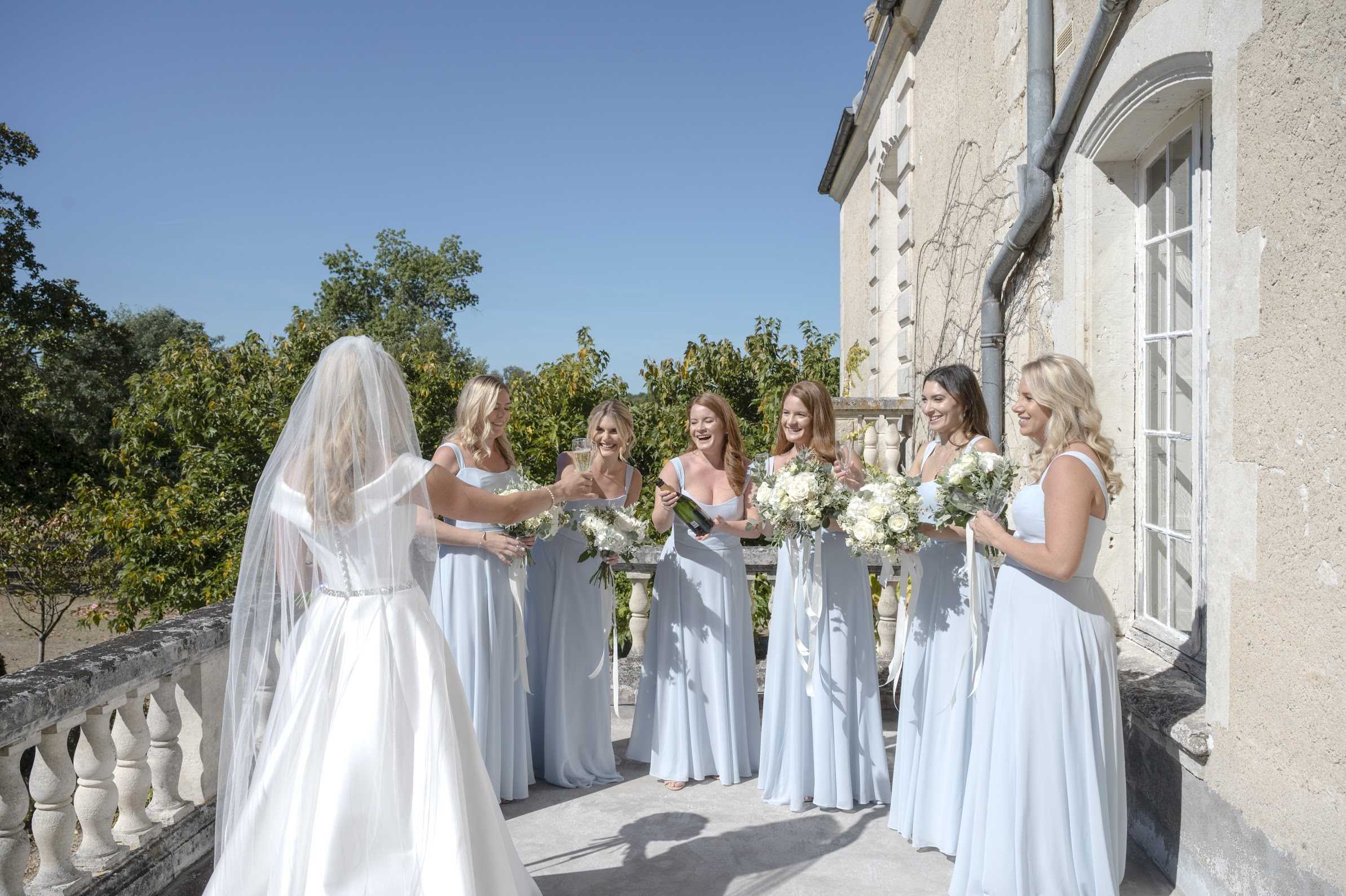 The bride and her five bridesmaids are toasting with champagne on an outdoor stone balustrade terrace of a French chateau, captured in a wide group shot. The bride, photographed from behind, wears a white satin ball gown with a crystal-embellished belt and a long cathedral-length veil. The five bridesmaids wear floor-length powder blue sleeveless dresses in a simple square-neckline style, and each holds a bouquet of ivory roses, white peonies, and silver-green dusty miller with trailing white ribbons. One bridesmaid is pouring champagne from a bottle while the group laughs and clinks glasses, creating a candid, celebratory moment. The classic French chateau facade with tall white-framed windows serves as the backdrop, contributing to the polished, classic aesthetic of the bridal party styling.