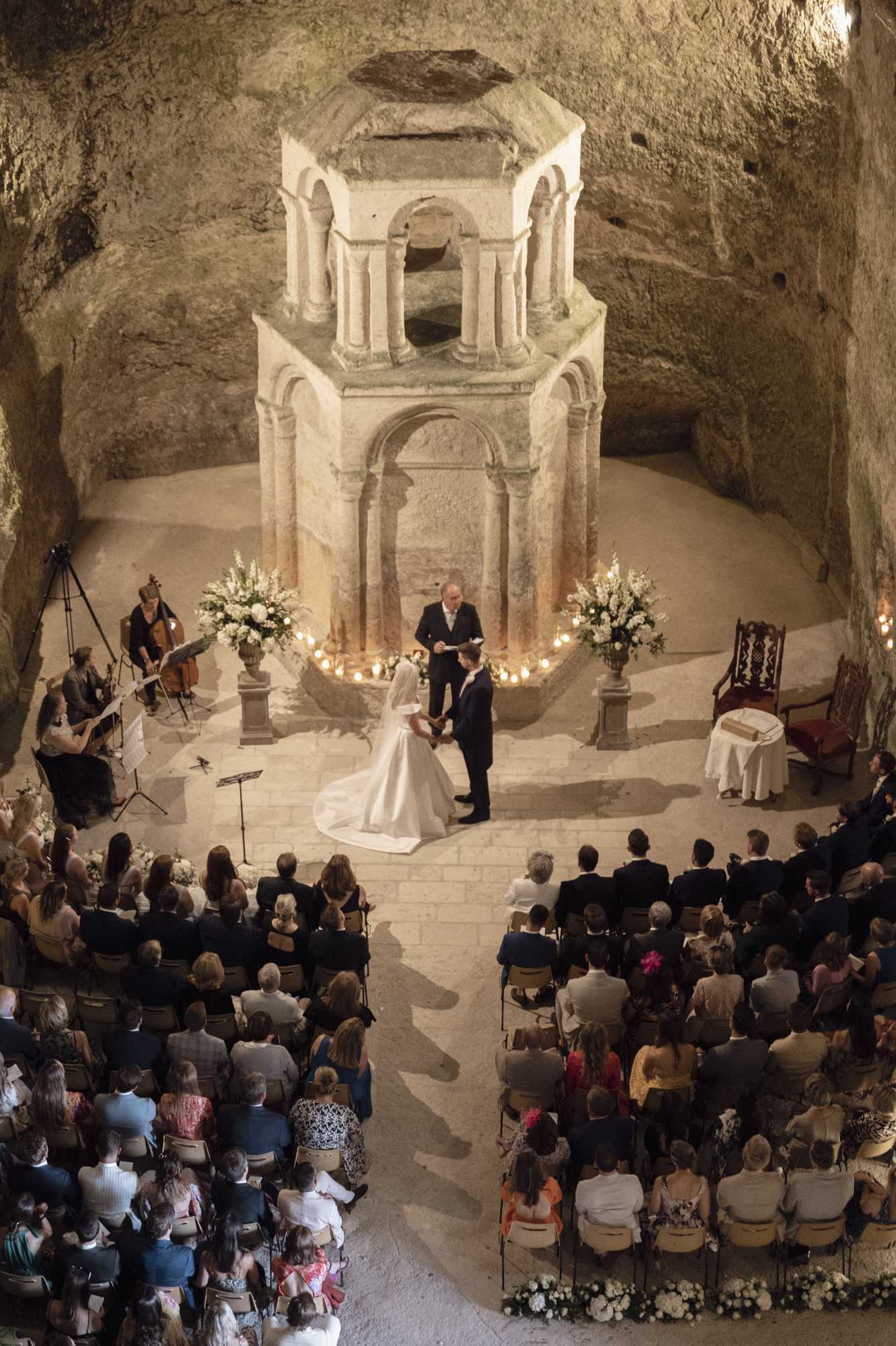 A wedding ceremony taking place inside an underground cave or troglodyte chapel, with the couple standing before an officiant at the base of a large octagonal stone structure with Romanesque arched columns carved directly into the rock. The bride wears a white ballgown with a long cathedral train, and the groom is in a dark suit. The altar area is lit by a semicircle of candles along the stone floor and flanked by two large urn arrangements of white flowers, likely white delphiniums or similar tall blooms. A string quartet performs to the left of the altar. Approximately 150 or more guests are seated in chairs arranged in two sections facing the altar. The wide overhead shot captures the full cave interior, the aisle, and the complete guest seating arrangement, giving a clear sense of the scale and architectural drama of the space. Potential venue feature image.