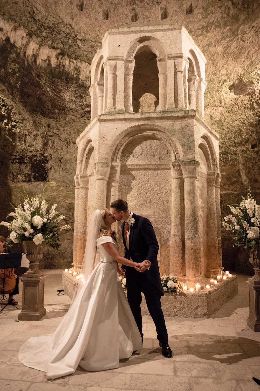A couple shares their first kiss during a wedding ceremony inside a cave or underground grotto, standing in front of a large ancient Romanesque stone structure with arched columns on two tiers. The bride wears an off-the-shoulder white satin ballgown with a cathedral-length veil and a thin embellished belt at the waist; the groom wears a navy suit with a patterned tie and a white buttonhole. The base of the stone monument is lined with glass votive candles and a low arrangement of white flowers, while two large stone urns flank the scene holding tall arrangements of white flowers — including what appear to be hydrangeas, stocks, and foliage — adding height on either side. The warm amber lighting from the candles and artificial lighting illuminates the carved rock walls of the cave interior, creating a dramatic, intimate atmosphere. This is a full-length portrait shot capturing both figures and the full architectural backdrop. Potential venue feature image.