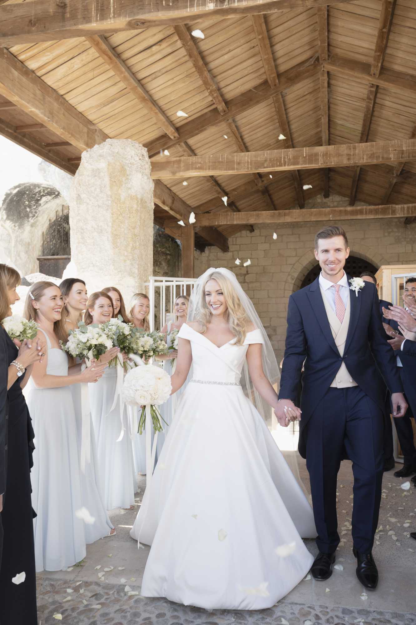 The bride and groom walk hand-in-hand during their post-ceremony recessional under an open-sided wooden-beamed structure attached to a stone chapel or church, with white flower petals being tossed into the air around them. The bride wears an off-the-shoulder white ballgown with a crystal-embellished belt and a cathedral-length veil, carrying a round bouquet of white peonies and ivory blooms with eucalyptus greenery. The groom is dressed in a navy suit with a champagne waistcoat and a coral/pink tie, wearing a white floral boutonniere. Approximately five to six bridesmaids line the left side wearing floor-length pale ice-blue dresses and holding white and green bouquets, while other guests clap and cheer on both sides; the overall styling palette is white, ivory, and pale blue with a classic, formal aesthetic. The shot is a medium-wide portrait-orientation image capturing the full couple and bridal party in natural daylight.