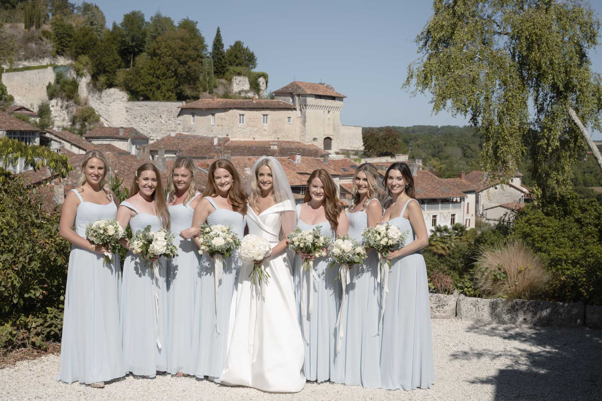 A bridal party portrait taken outdoors on a gravel terrace with a historic French village and stone château visible in the background. The bride stands at center wearing an off-the-shoulder structured white ballgown with a cathedral-length veil, while seven bridesmaids flank her in floor-length ice blue chiffon dresses with square necklines and varying strap styles. All eight women hold rounded bouquets of ivory garden roses, white peonies, and eucalyptus with trailing ivory ribbon. The overall color palette is white and pale blue with soft greenery accents, giving the group a classic, clean aesthetic. This is a wide group portrait shot in natural daylight.