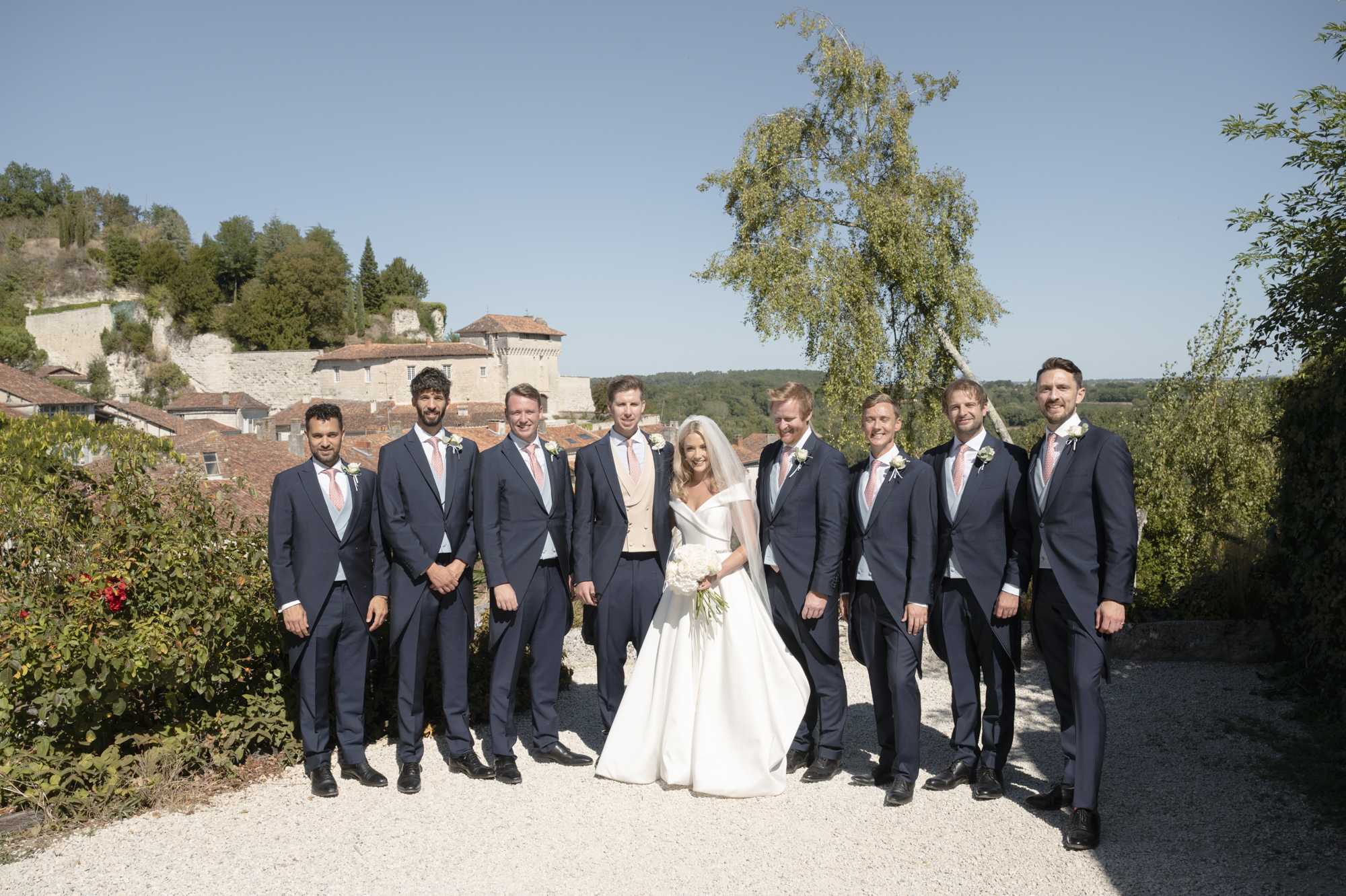 A group portrait taken outdoors on a gravel path, featuring the bride surrounded by eight groomsmen against a backdrop of a French village with historic stone buildings and terracotta rooftops. The bride wears an off-the-shoulder white ballgown with a cathedral-length veil and carries a bouquet of white peonies and ivory blooms. The groom stands at center in a champagne waistcoat with navy trousers, while the seven groomsmen all wear matching navy suits with blush pink ties and white floral buttonholes. The classic, formal styling is consistent across the group, and the wide-shot composition captures all nine subjects at full length in bright afternoon daylight.