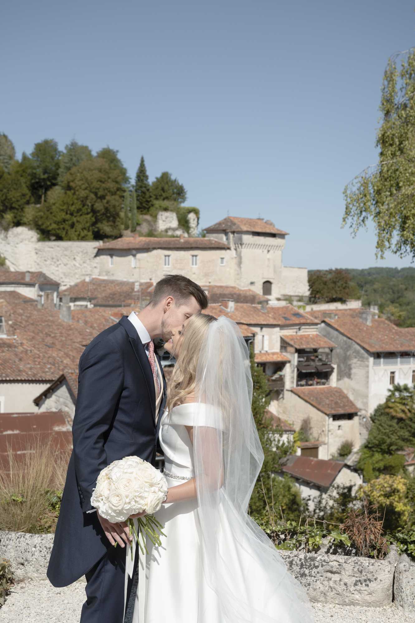 A couple portrait taken outdoors at a elevated viewpoint overlooking a traditional French village with terracotta-roofed stone buildings and a castle or fortified manor in the background. The groom wears a navy suit with a blush pink tie and leans in to kiss the bride on the forehead. The bride wears an off-the-shoulder white satin gown with a crystal-embellished waist detail and a long tulle veil, and holds a tightly rounded bouquet of cream roses with white ribbon streamers. The composition is a mid-range portrait shot with the village rooftops and hillside architecture providing a distinctly French backdrop.