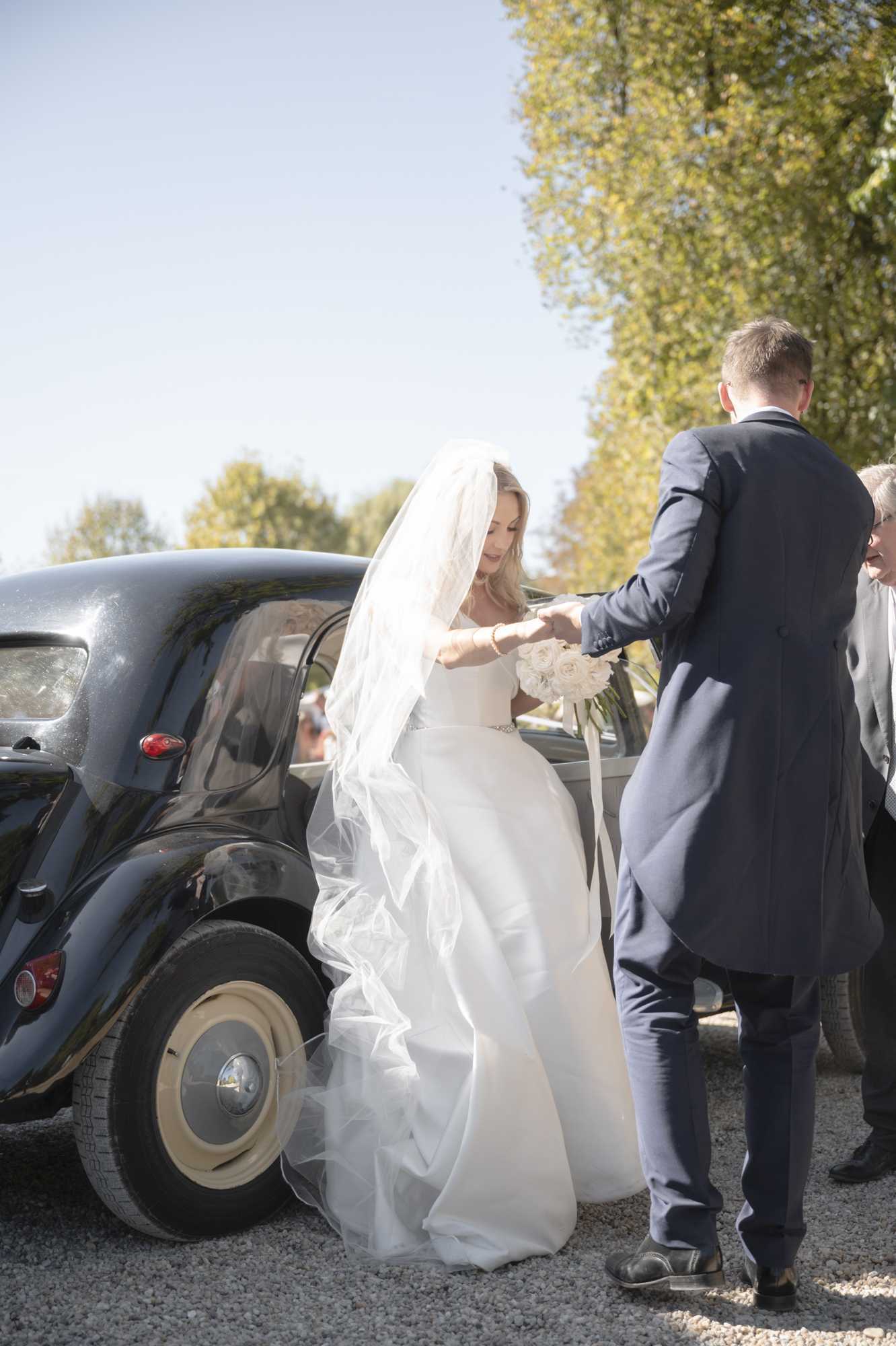 A bride is being helped out of a vintage dark grey/black classic car, likely a Citroën 2CV or similar French vintage vehicle, on a gravel surface outdoors. A man in a navy blue suit is assisting her by holding her hand as she steps out, while another guest in grey stands nearby. The bride wears a white strapless ball gown with a full tulle skirt and a long cathedral-length tulle veil, and carries a compact round bouquet of white roses. The setting and vintage car choice suggest a classic, polished wedding style. This is a mid-range portrait shot taken in bright natural daylight.