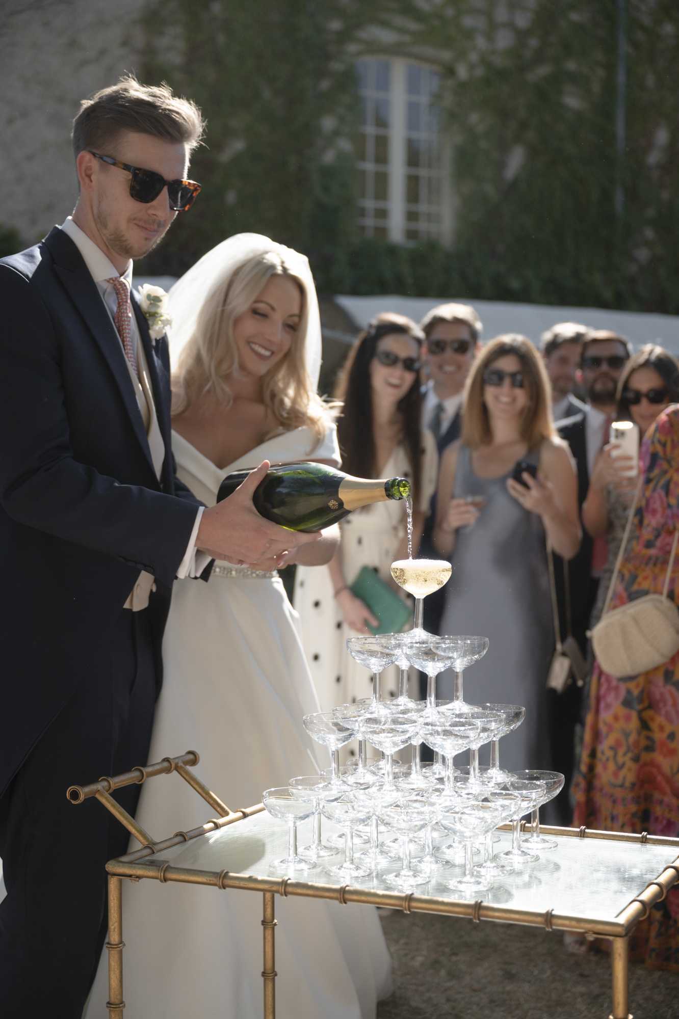 The couple is pouring champagne into a coupe glass tower during an outdoor cocktail hour, with the groom in a navy suit, salmon-pink tie, and dark sunglasses holding a large champagne bottle, and the bride in an off-the-shoulder white gown with a beaded waist detail and a veil. The champagne tower is arranged on a gold bamboo-style bar cart with a mirrored top, and the champagne is visibly cascading down through the stacked coupe glasses. Approximately six to eight guests are gathered behind the couple, smiling and photographing the moment on their phones, dressed in formal attire including a grey midi dress and a floral printed gown. The setting is an outdoor courtyard in front of what appears to be a stone château building, and the portrait-oriented medium shot captures both the couple and the reacting guests in natural sunlight.