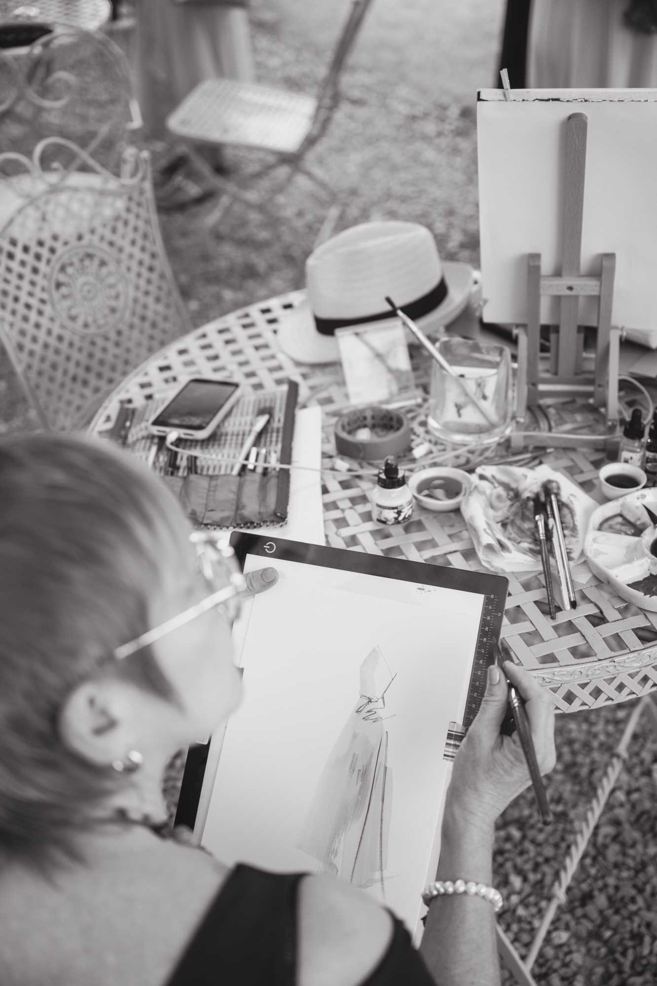 A black-and-white overhead close-up shot of a live wedding illustrator at work during what appears to be a cocktail hour or reception, seated outdoors at a lattice-patterned bistro table. The artist, a woman wearing a beaded bracelet, holds a paintbrush in each hand and is sketching a figure in a wedding gown on paper placed over an illuminated light pad. The round table is covered with art supplies including ink bottles, small paint pots, a glass jar holding brushes, a smartphone, a notebook, and a panama-style hat with a dark band. A small wooden easel holding a blank canvas is visible in the upper right corner, and ornate metal bistro chairs are visible in the background.
