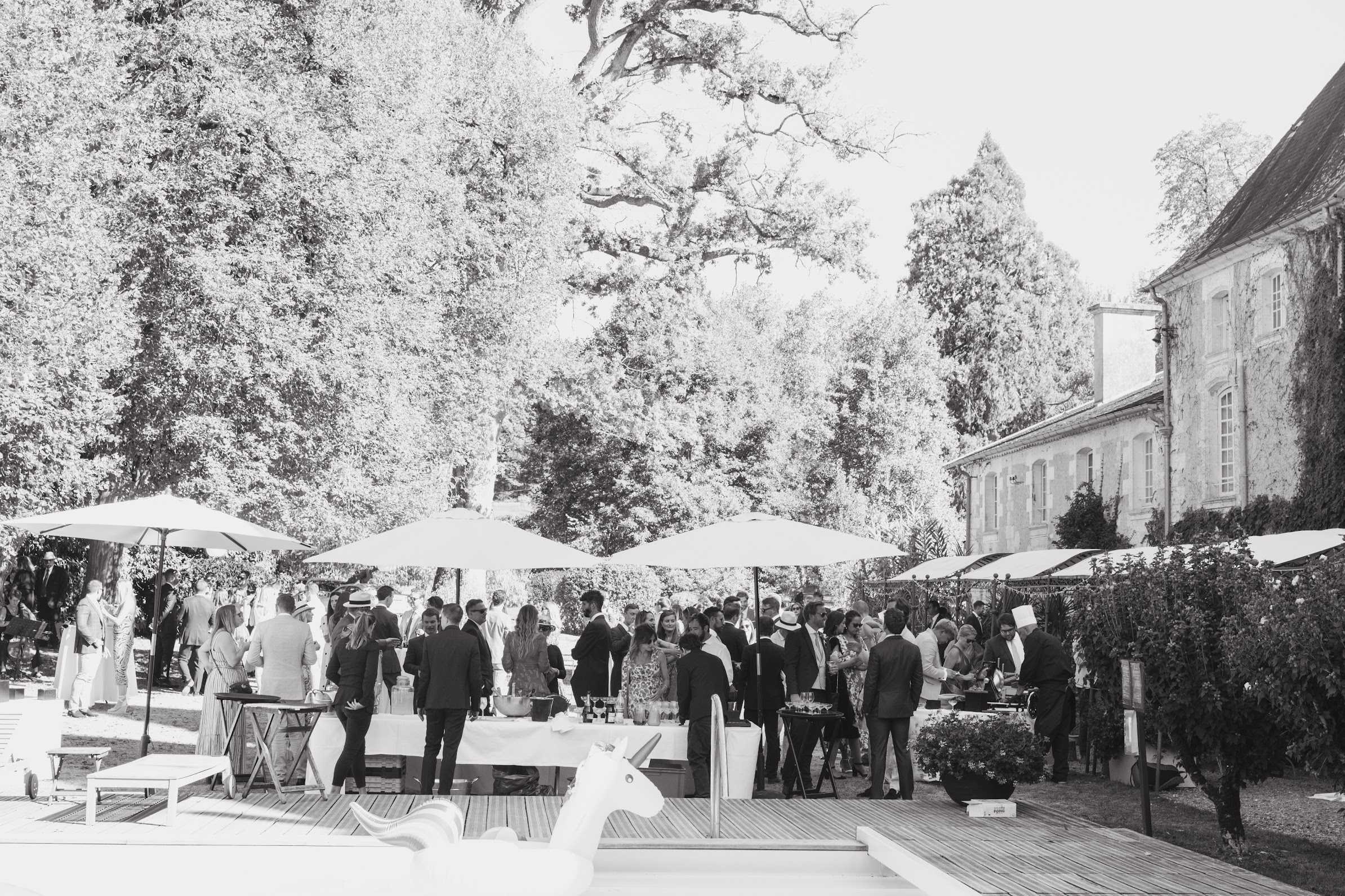 This black-and-white wide shot captures an outdoor cocktail hour with approximately 50–60 guests mingling on a wooden pool deck beside what appears to be a French chateau, visible on the right edge of the frame in classic stone construction. Several large market umbrellas are positioned across the deck to provide shade, with catering stations and beverage tables set up among the crowd; a chef in a white toque is visible near one of the food stations on the right. Guests are dressed in formal and smart-casual attire, with several men in dark suits and women in dresses, and at least one guest wearing a wide-brim sun hat. A white inflatable unicorn float is visible in the foreground at the pool's edge, adding a playful, informal touch to the classic chateau setting. Potential venue feature image.