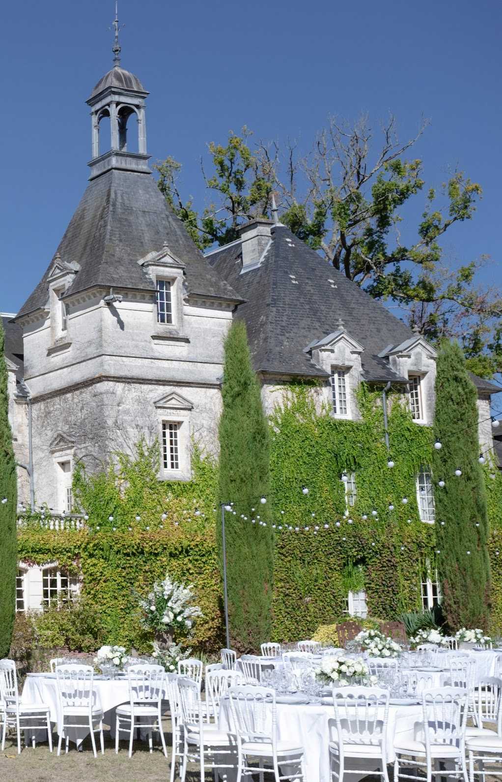 An outdoor wedding reception is set up in the grounds of a French chateau, with multiple round tables dressed in white linen and surrounded by white Napoleon/Chiavari chairs. The tables are decorated with white and ivory floral centerpieces featuring roses and greenery, and strings of globe lights are draped across the space in front of the building. The chateau itself is a classic French stone manor with slate-roofed conical towers, a belltower cupola, and ivy-covered facades, giving the setting a classic formal aesthetic. This is a wide establishing shot taken from ground level showing the full venue exterior alongside the reception layout. Potential venue feature image.