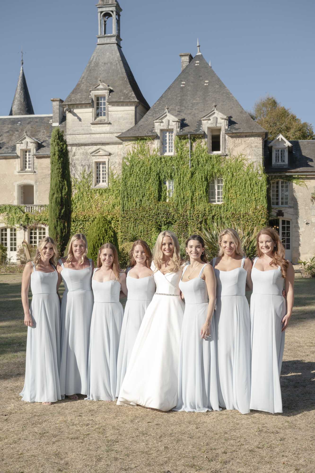 A bridal party portrait taken outdoors on the grounds of a French chateau, with the bride standing at the center flanked by seven bridesmaids arranged in a line. The bride wears a white satin ballgown with a V-neckline and a crystal-embellished belt, while all seven bridesmaids wear floor-length ice blue sleeveless dresses with square necklines and a flowy chiffon skirt. The group is posed facing the camera in a wide portrait shot, smiling, with the multi-turreted limestone chateau and its ivy-covered facade visible behind them. Potential venue feature image.