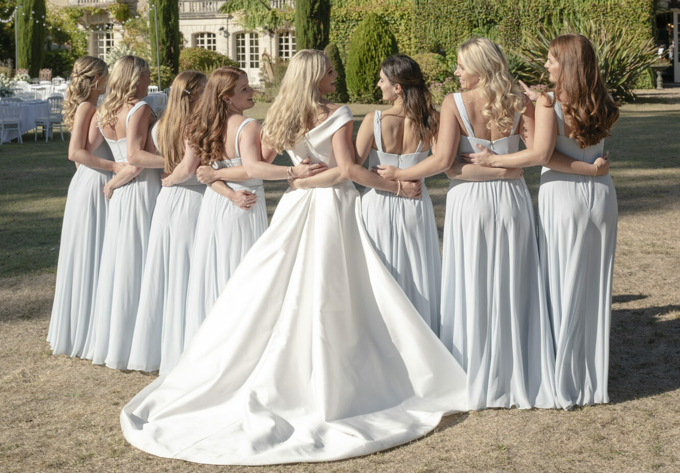 A bridal party portrait taken outdoors on the grounds of a French chateau, showing the bride surrounded by seven bridesmaids, all photographed from behind with arms linked around each other's waists. The bride wears a white satin ballgown with a structured off-the-shoulder neckline and a long cathedral-length train, while the bridesmaids wear matching floor-length pale ice-blue chiffon gowns with single-shoulder straps. The group is laughing and chatting animatedly, creating a candid feel within a posed group shot. In the background, ivy-covered chateau architecture is visible along with tall cypress trees, manicured hedging, and white-linened reception tables set with ghost chairs visible to the left. The shot is a wide, medium-distance portrait taken at ground level.