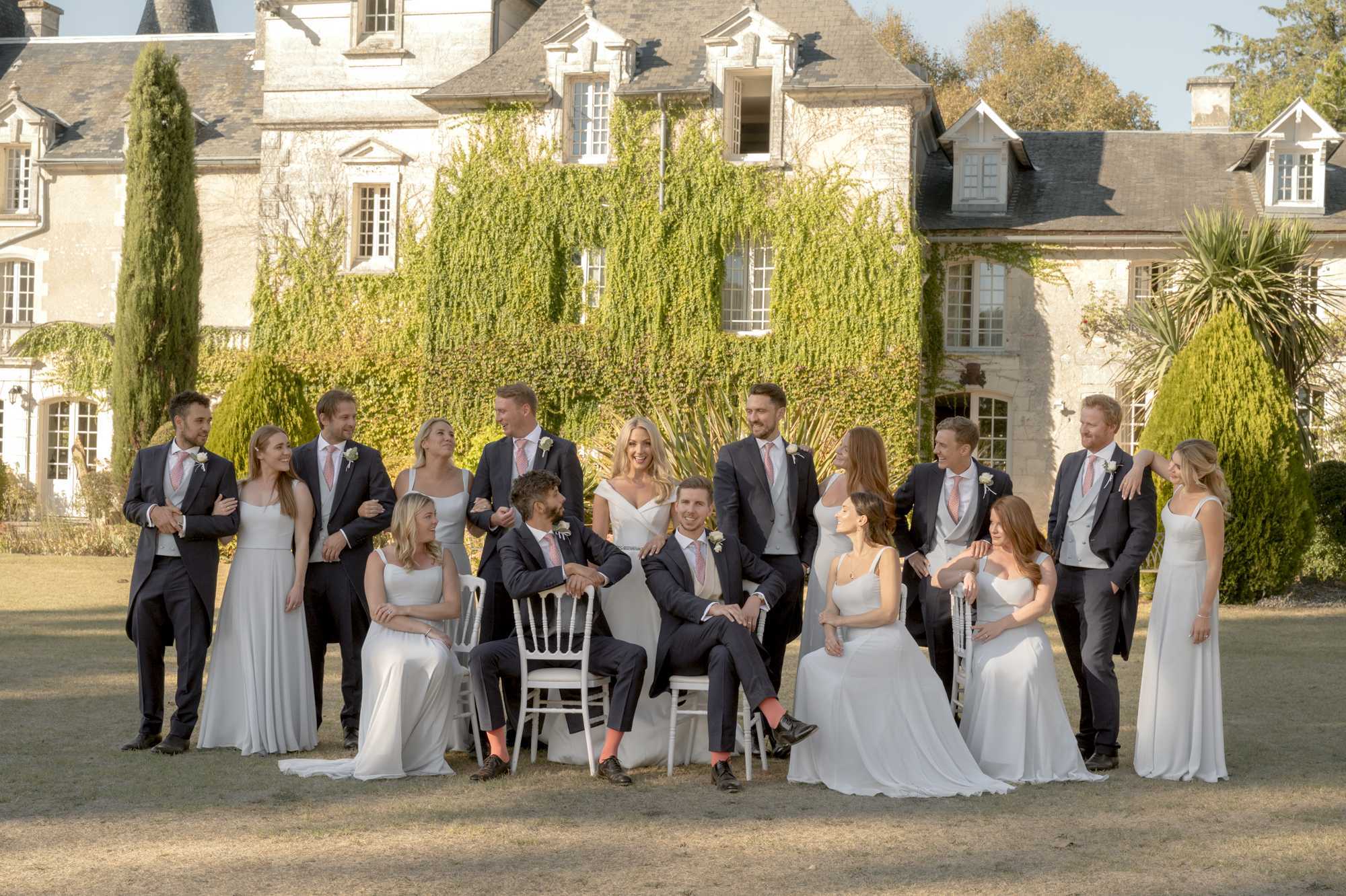 A bridal party group portrait taken outdoors on the grounds of a French chateau, with the ivy-covered stone facade of the building prominently visible in the background. The group of approximately 16 people includes the bride in an off-the-shoulder white fitted gown and the groom in a navy suit with a coral-pink tie and matching coral socks, both seated on white Chiavari chairs at the center. Bridesmaids wear floor-length light grey/silver chiffon dresses, while groomsmen are dressed in charcoal morning suits with light grey waistcoats and coral-pink ties with small white buttonhole flowers. The composition is a wide shot with some members standing and others seated, arranged in a relaxed, candid-style pose with people interacting and laughing, shot in warm afternoon sunlight. Potential venue feature image.