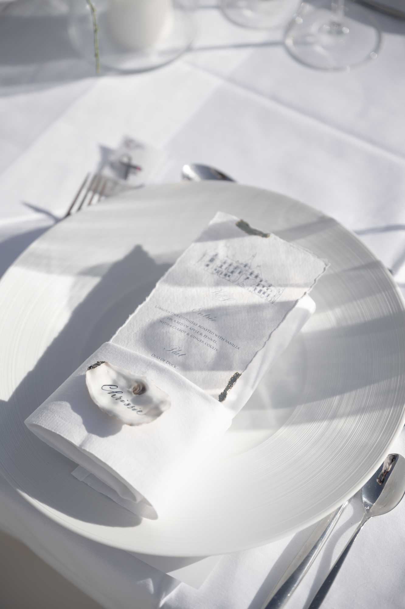 Close-up detail shot of a wedding reception place setting featuring an all-white palette. A white round plate sits on a white linen tablecloth, holding a folded white linen napkin topped with a deckle-edged menu card printed in fine script lettering — menu items partially legible include references to duck and carrot with vanilla and fennel. A small oyster shell with calligraphy reading 'Christine' serves as the place card, and two small agate or stone slices act as decorative accents on the napkin. Silver cutlery including a fork, spoon, and knife is arranged around the plate, with clear glassware and a small white ceramic vessel visible in the background. The styling is minimal and modern with a monochromatic white-on-white aesthetic, shot at an angle in bright natural light that casts soft shadows across the linen.