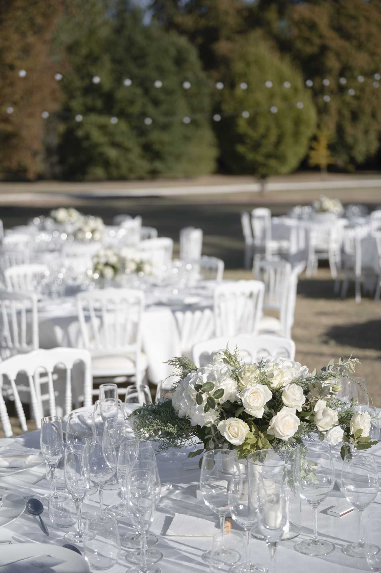 An outdoor wedding reception setup photographed in a medium close-up shot, focusing on a round guest table in the foreground with multiple additional tables visible and slightly out of focus behind it. The decor follows a strict all-white and green palette: tables are dressed in white linens and surrounded by white Napoléon-style chairs, with low centerpieces of ivory roses, white hydrangeas, eucalyptus, and feathery green foliage arranged in clear glass vessels. Each place setting includes multiple crystal wine and champagne glasses, white plates, and silver cutlery. Tall hurricane candle holders with white candles are also visible on the tables. String lights with globe bulbs are strung overhead across the outdoor terrace space, and what appears to be a reflecting pool or water feature is visible in the background. The overall styling is classic and clean, with a modern French formal aesthetic.