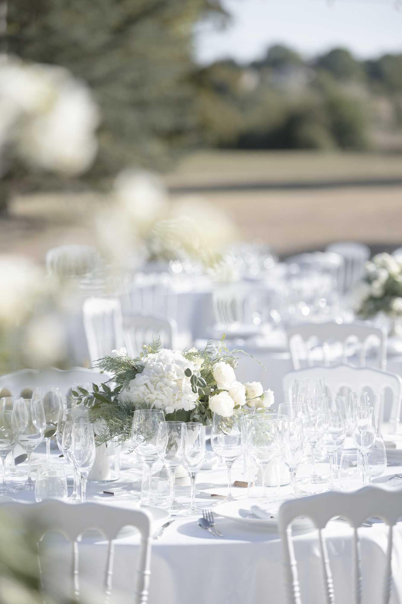 An outdoor wedding reception table setup shot in natural daylight, showing multiple round tables dressed in white floor-length linens with white Napoléon chairs. The closest table features a low centerpiece of white hydrangeas, white garden roses, eucalyptus, and feathery green fern foliage in a white vessel, with additional matching centerpieces visible on tables receding into the background. Each place setting includes multiple clear crystal wine and water glasses, silver cutlery, white folded napkins, and small place cards. The overall decor palette is an all-white and green scheme with a classic, clean aesthetic. The shot is a medium close-up taken at table level with shallow depth of field, with blurred white floral arrangements in the foreground framing the composition.