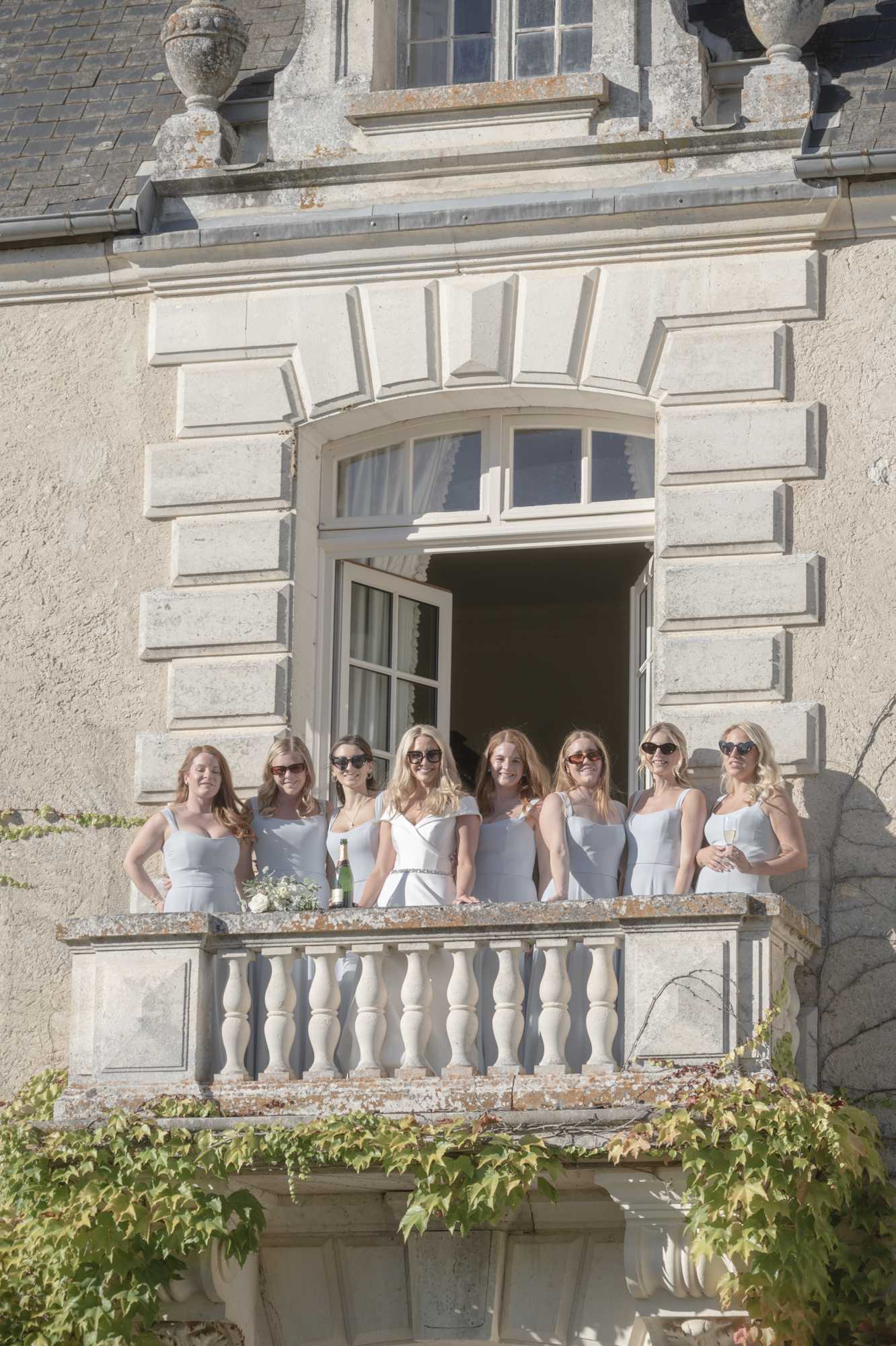 A bride and her seven bridesmaids pose together on a stone balcony of a French chateau during what appears to be a getting-ready or pre-ceremony moment. The bride wears a white off-the-shoulder fitted dress with a belted waist, while the bridesmaids are dressed in matching pale blue-grey sleeveless fitted dresses; most of the group wear sunglasses. A champagne bottle and a small white bouquet rest on the balcony railing, and one bridesmaid holds a champagne flute. The shot is a wide exterior portrait framed by the chateau's classical limestone facade with arched French windows and a balustrade railing. Potential venue feature image.
