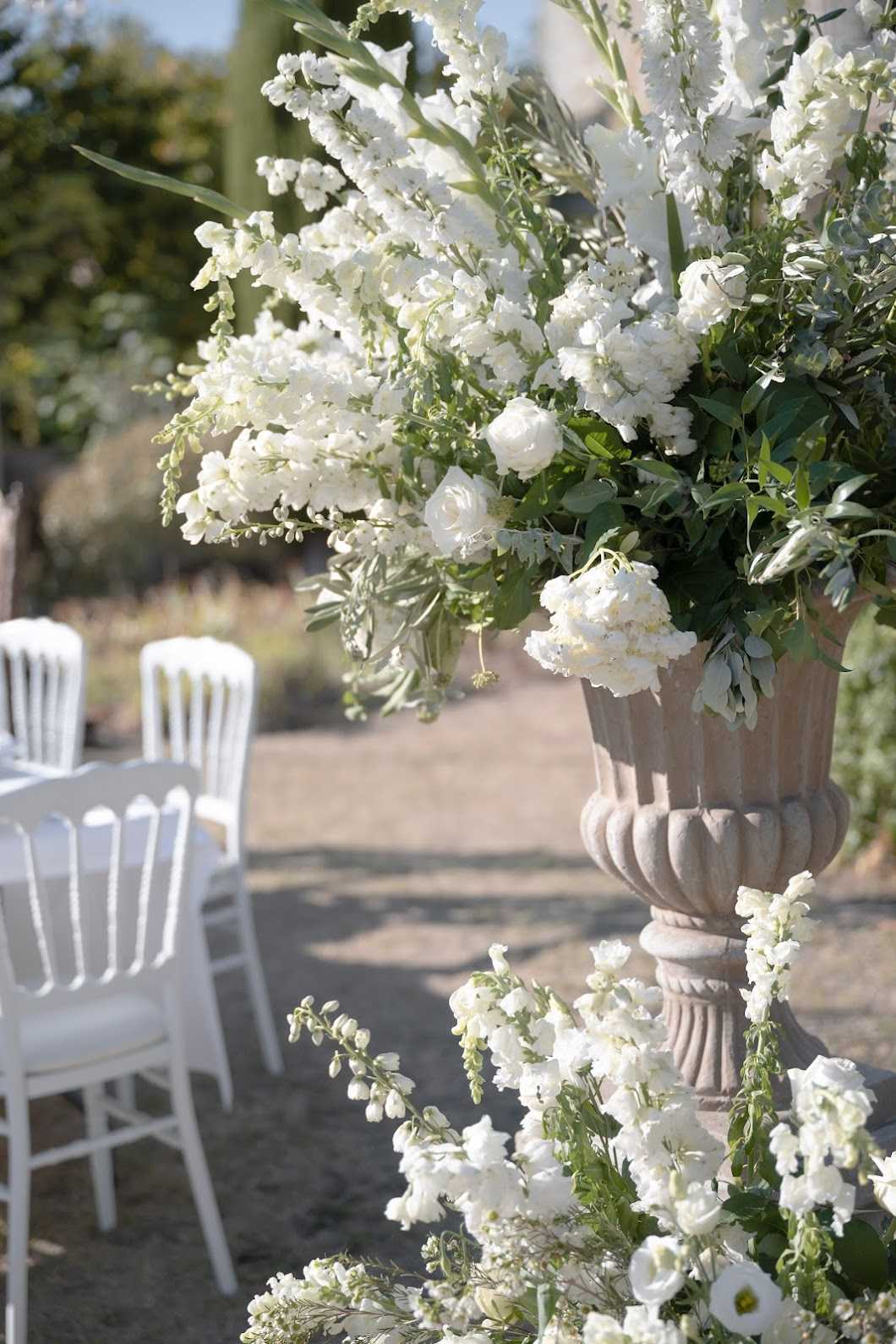 Close-up detail shot of an outdoor wedding ceremony setup featuring a large stone urn overflowing with an all-white floral arrangement composed of white delphiniums, white roses, white hydrangeas, white lisianthus, and trailing white snapdragons mixed with olive branch foliage and green leaves. The stone urn has a classical pedestal form in a warm beige-grey tone. Additional white blooms cascade down the base of the urn in the foreground. In the soft-focus background, white Napoleon (Chiavari-style) chairs are arranged in rows on a gravel surface, indicating an outdoor ceremony aisle. The overall decor palette is a strict white and green combination with a classic, garden-inspired styling.