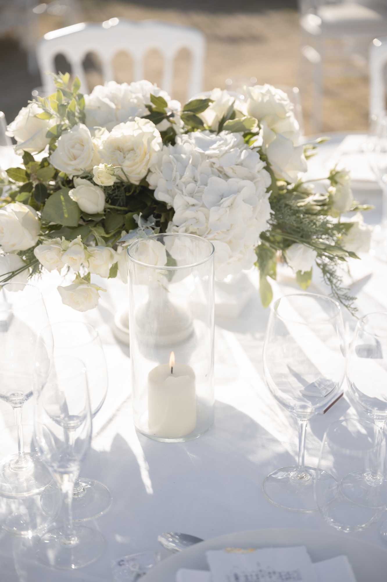 Close-up detail shot of an outdoor wedding reception table set in an all-white color palette. The centerpiece features a low, lush arrangement of white roses, white hydrangeas, and green foliage, paired with a tall glass hurricane vase holding a lit white pillar candle. The round table is dressed in a white linen tablecloth and set with clear crystal wine glasses, champagne flutes, and white folded napkins, with place cards visible at the edges. White Napoléon chairs are visible in the soft-focus background, suggesting a classic, clean aesthetic for the reception setting.